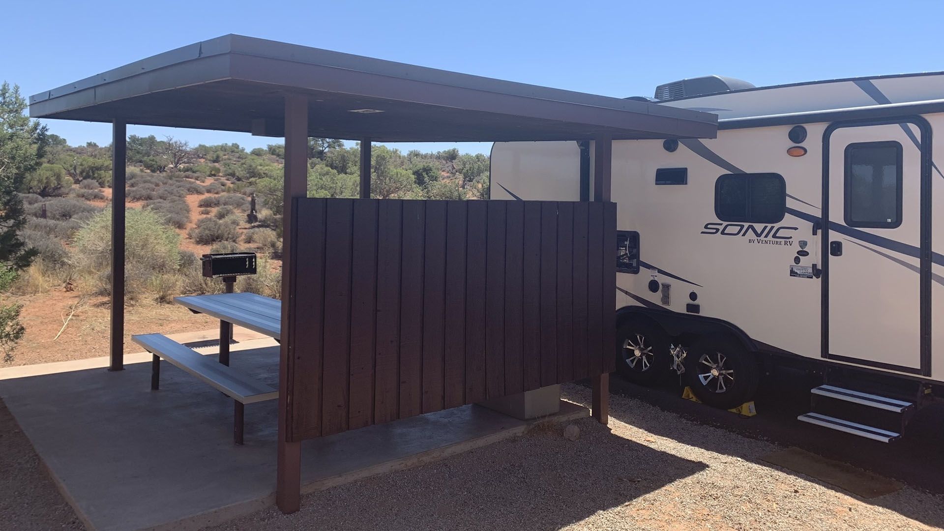RV parked next to a picnic area with a table, grill, and windbreak.
