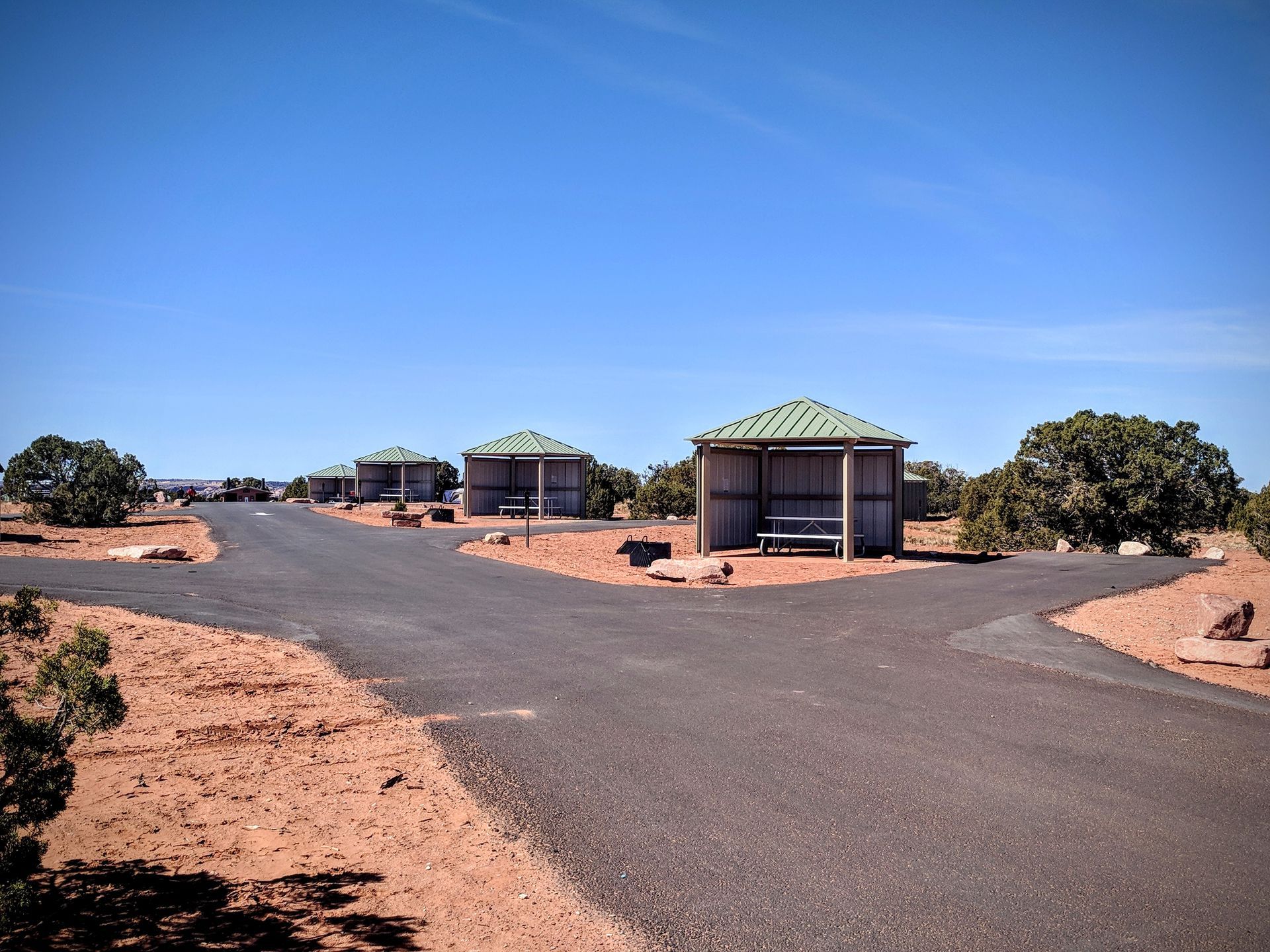 Three open-air shelters with green roofs line a paved road on a desert landscape under a blue sky.