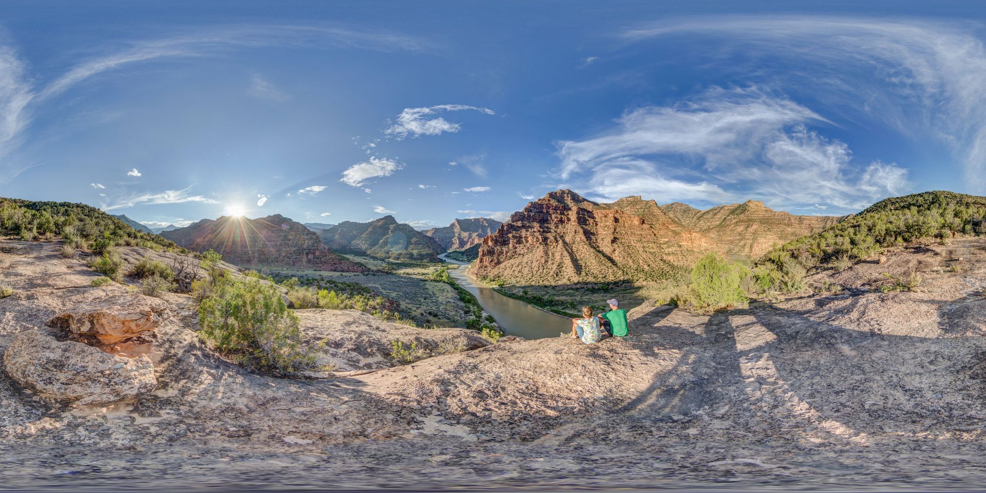 A father and son sit at the edge of a cliff with a dramatic canyon and river winding into the distance at sunrise.