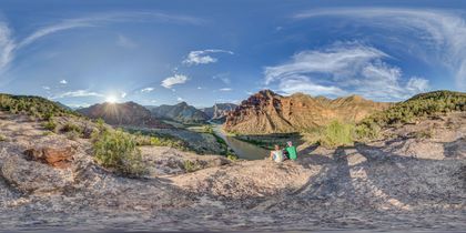 A father and son sit at the edge of a cliff with a dramatic canyon and river winding into the distance at sunrise.