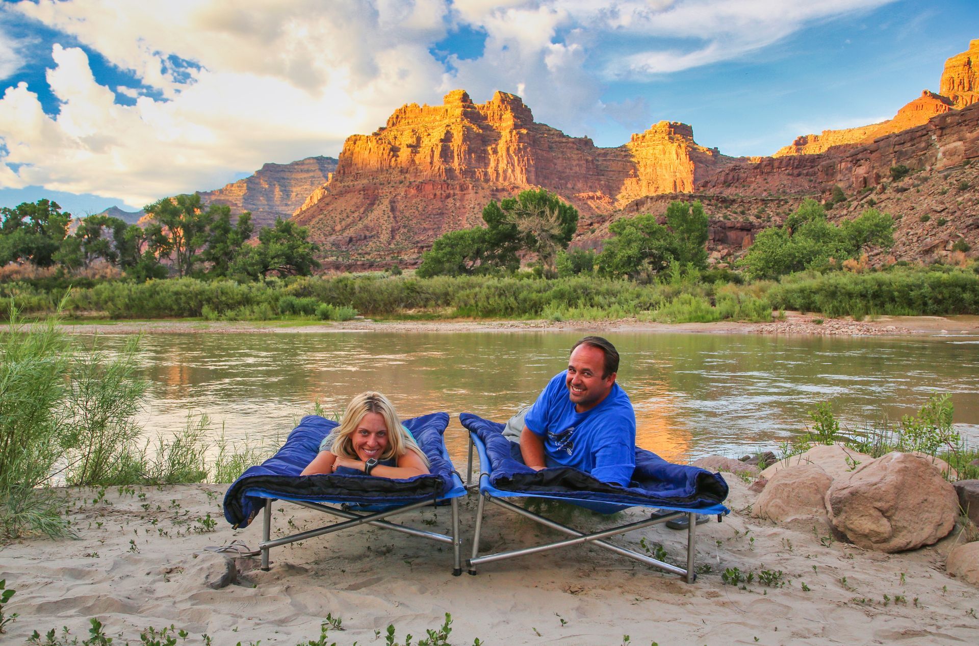 A couple lying on cots on a sandy riverbank with a Cataract Canyon backdrop at sunset.