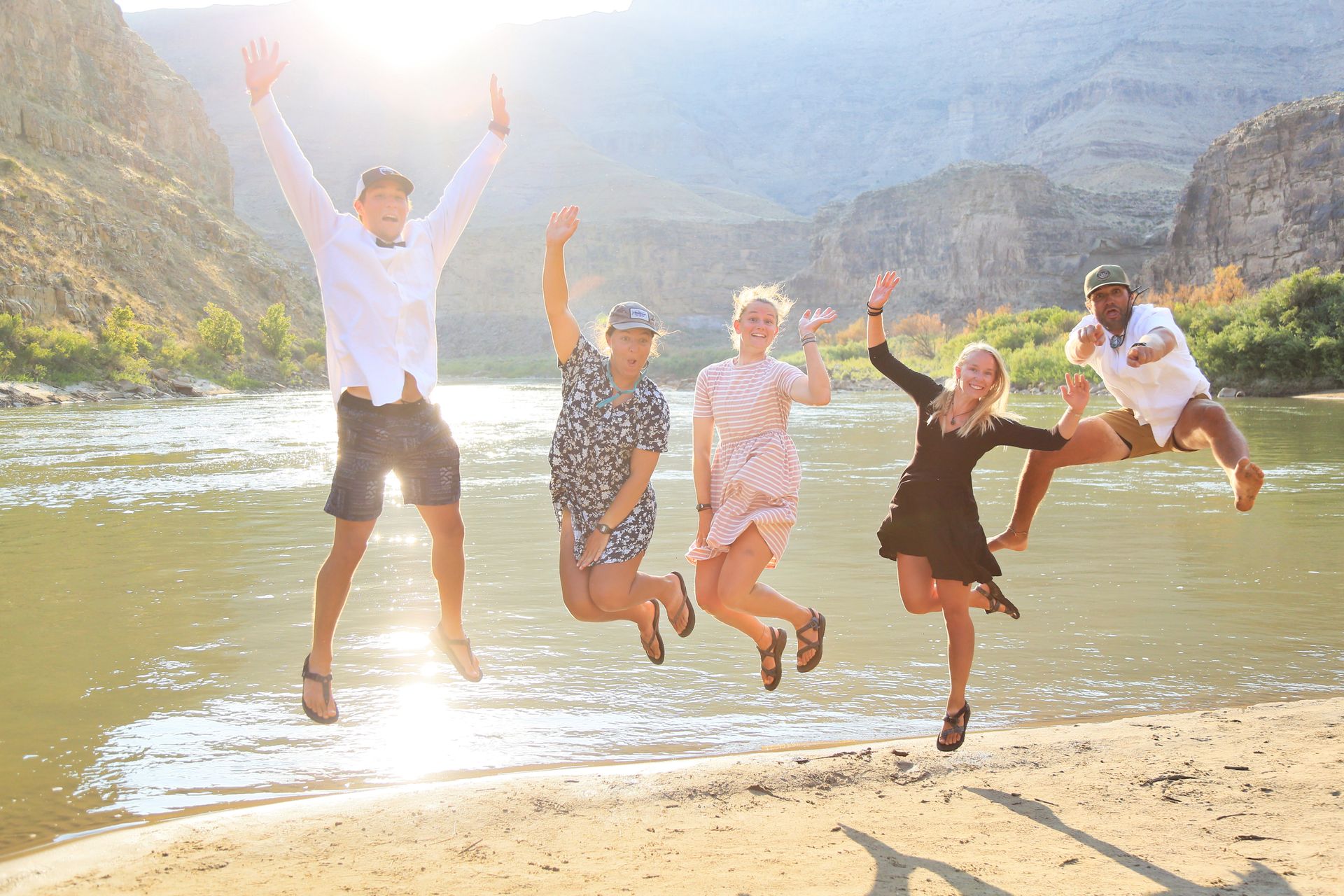 Five people jumping for joy on a sandy riverbank, mountains in background, sunny day.