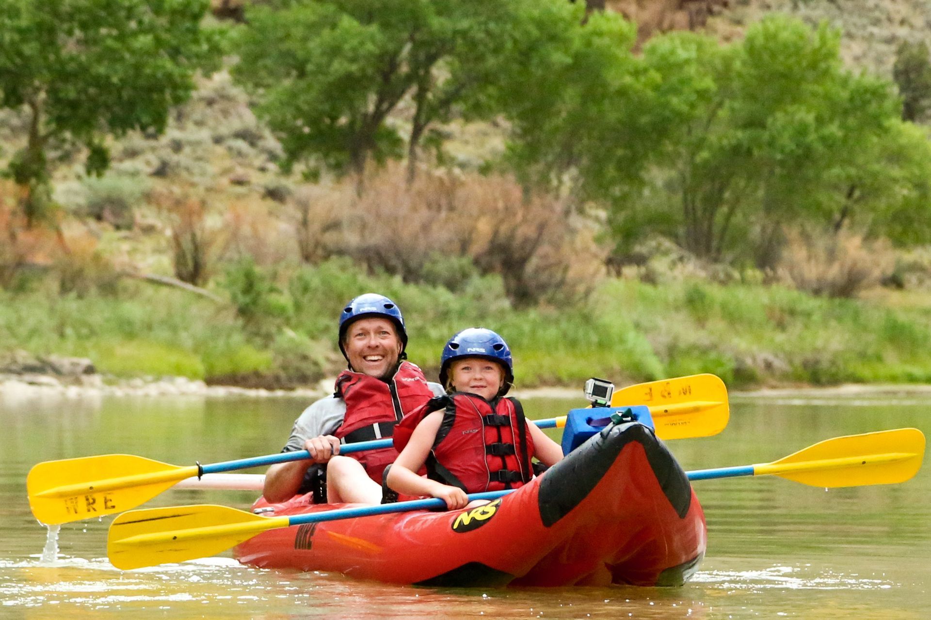 Woman and child kayaking on a river wearing life vests and helmets. Green trees in background.