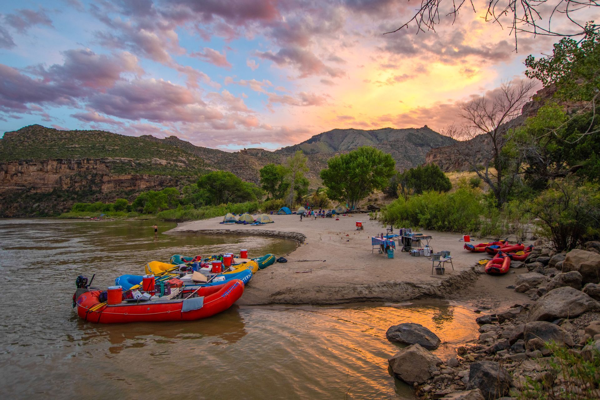 Rafts and campers on a sandy riverbank at sunset, mountains in the background.