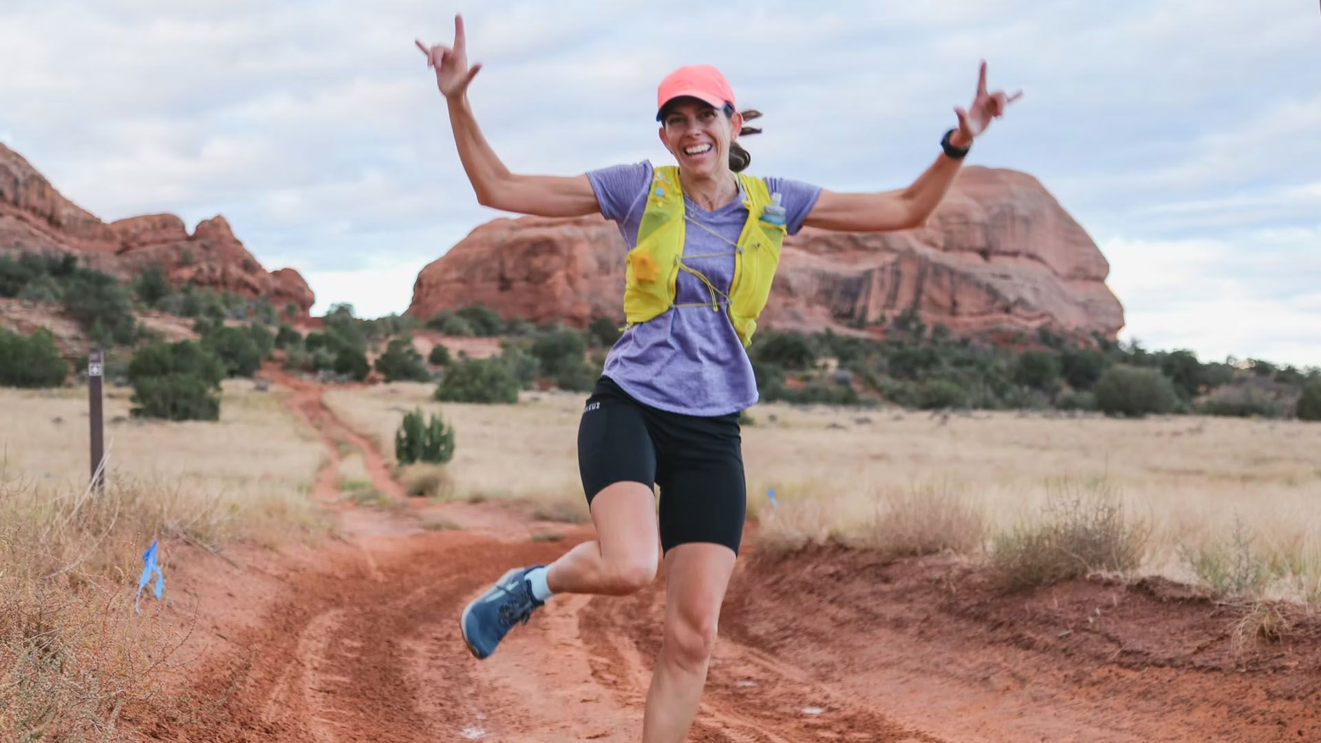 Woman trail running, arms raised in victory, on a dirt path in a desert landscape.