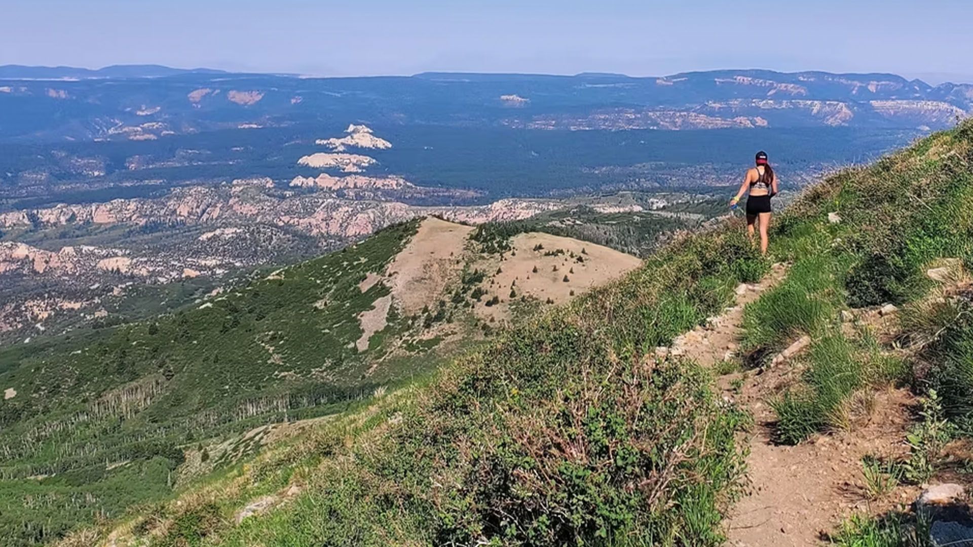 Woman runs along a mountain trail, vast green landscape and blue sky in the background.