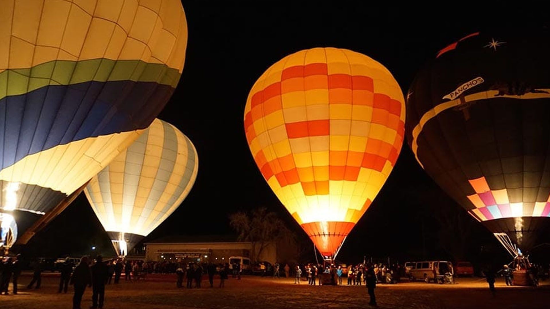 Hot air balloons at night, lit up with vibrant colors, people watching.