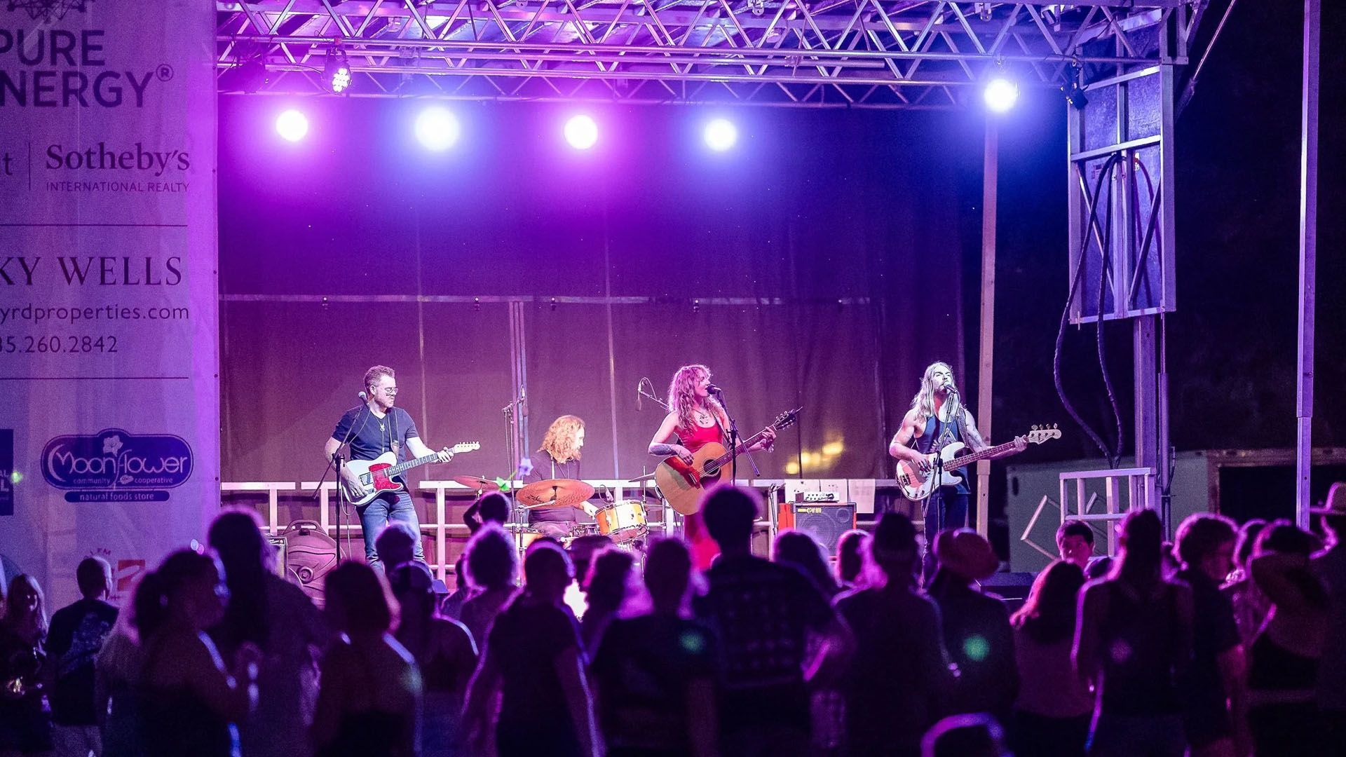 Band performing on outdoor stage with purple lighting, in front of a crowd.