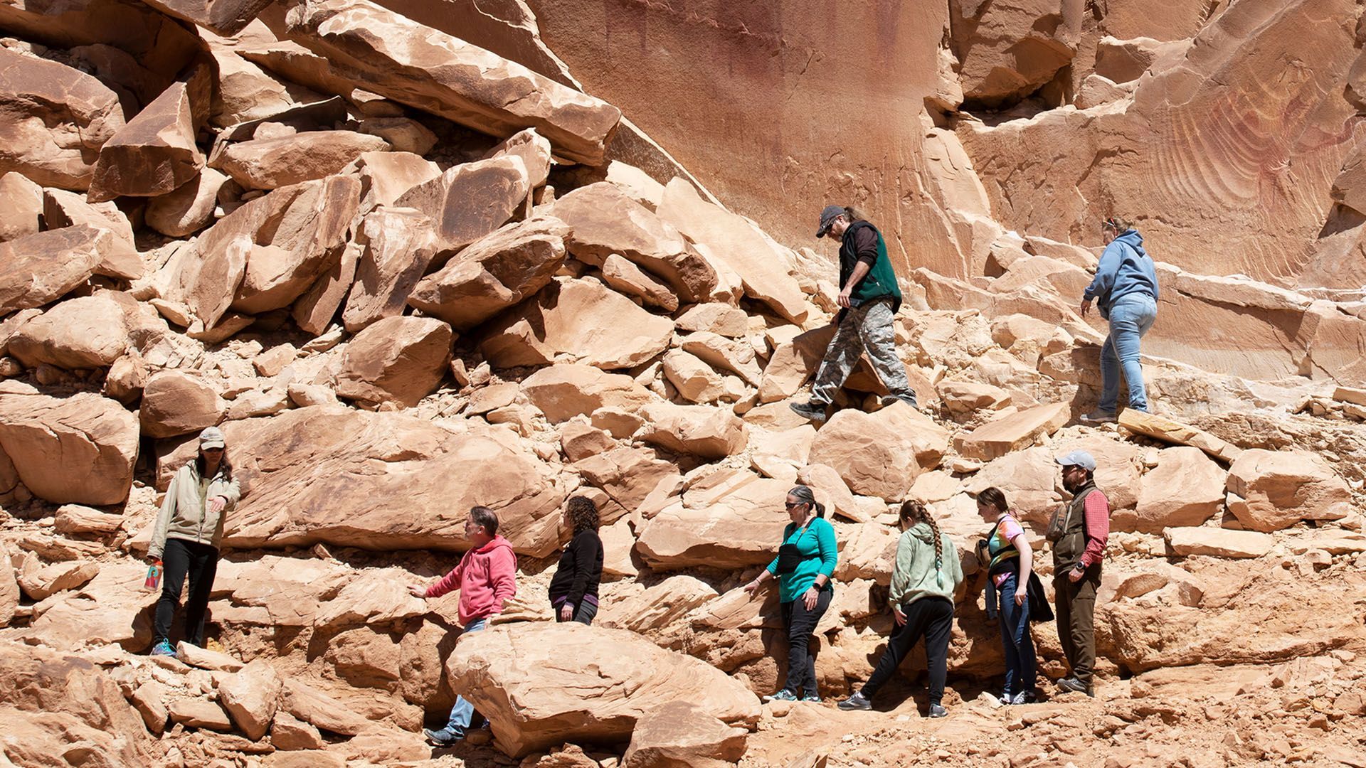 Group of people exploring a reddish-brown rock formation. They climb and examine the textured surface in a desert landscape.