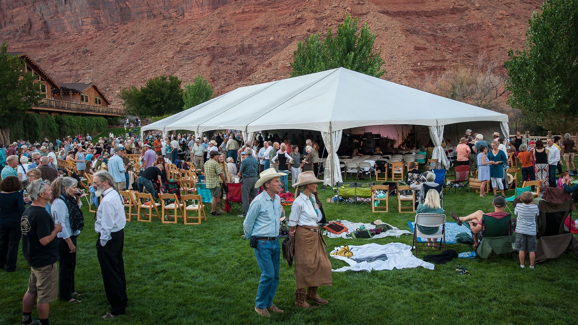 Large outdoor event under a white tent on a grassy area; red rock backdrop, people mingling.