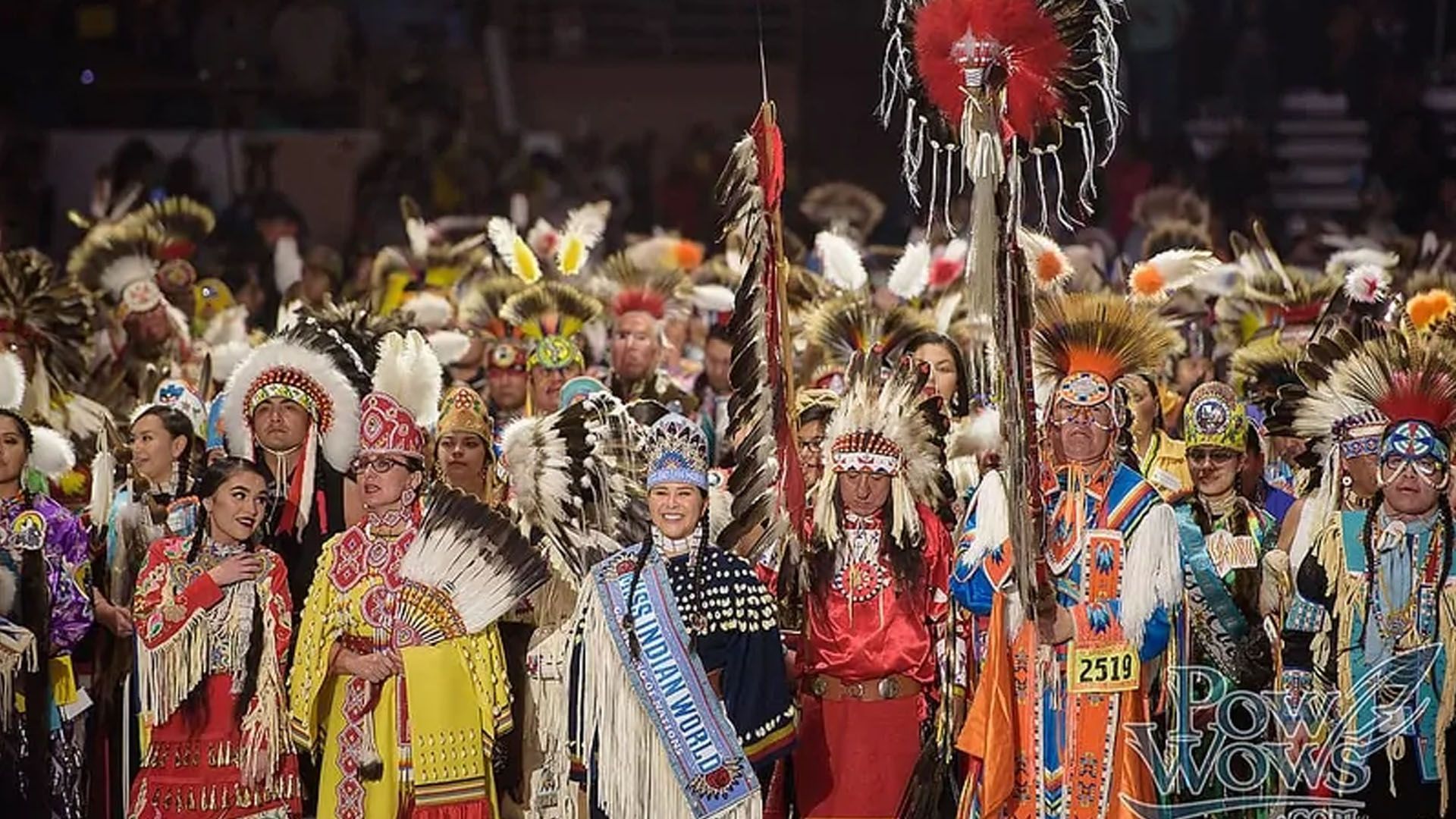 People in vibrant Native American regalia at a powwow. Feathers, beads, and color abound, indoors.