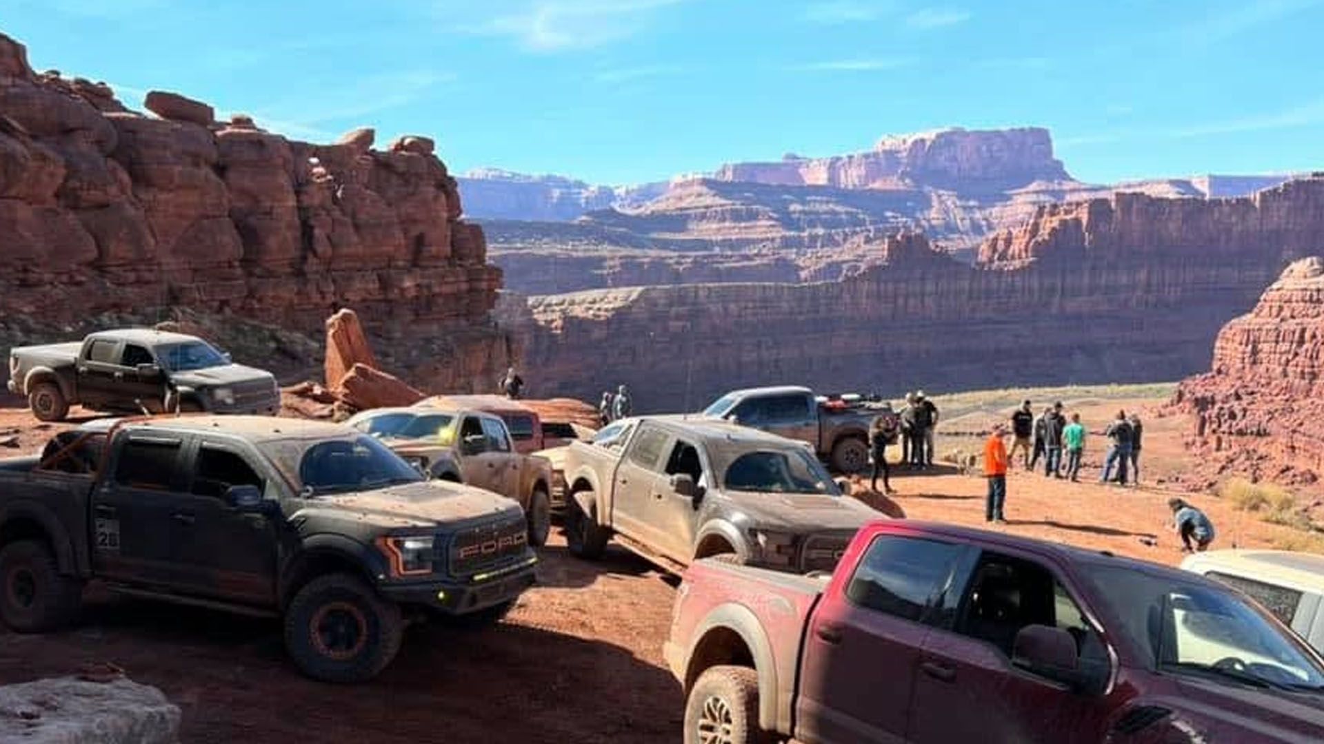 Off-road trucks parked on a red-rock mountain overlook; people standing nearby, blue sky.