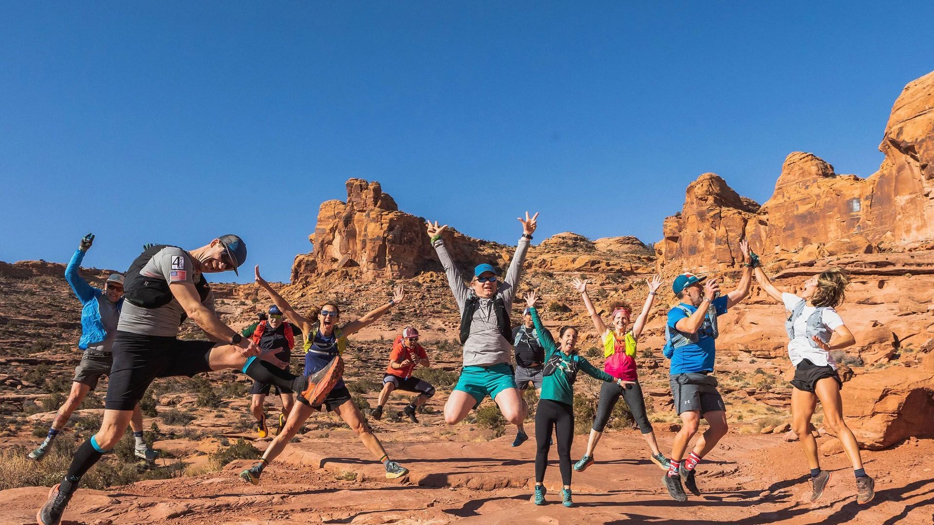Group of trail runners jumping joyfully in a red rock canyon under a blue sky.