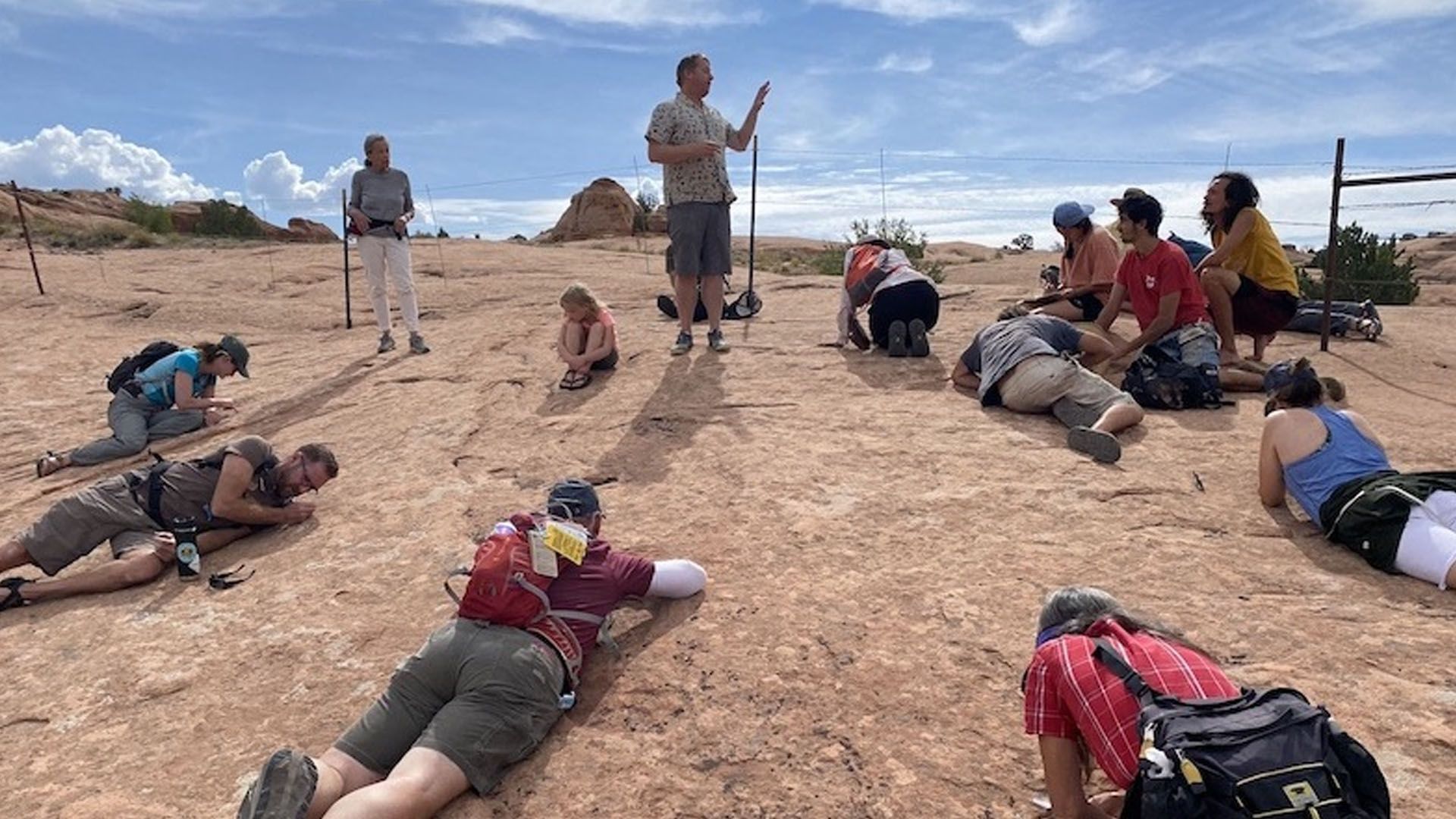 Group on red rock terrain, listening to a guide. Some people are lying down, looking at the ground.
