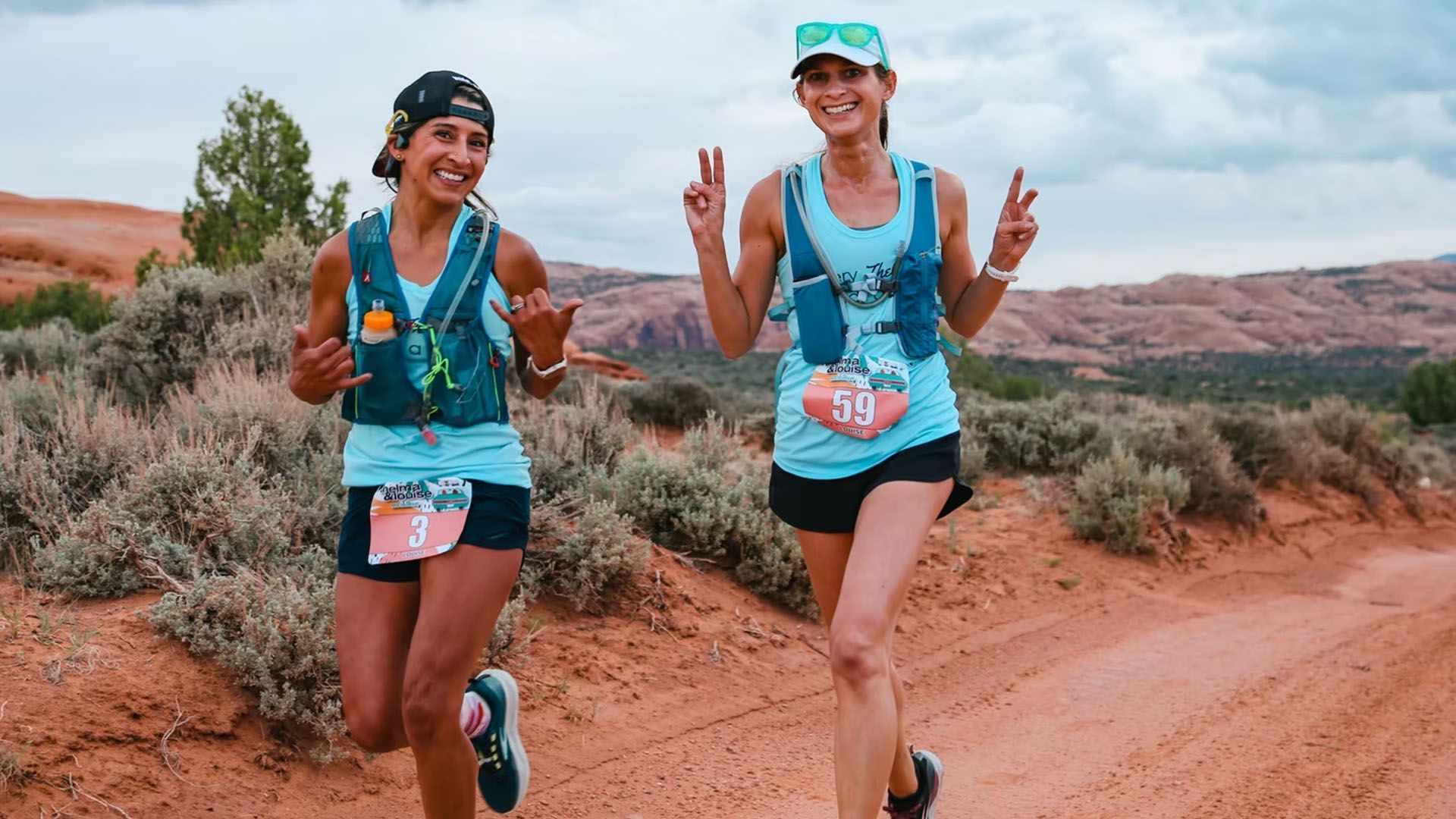 Two women running on a red dirt road in the desert, smiling and giving peace signs.
