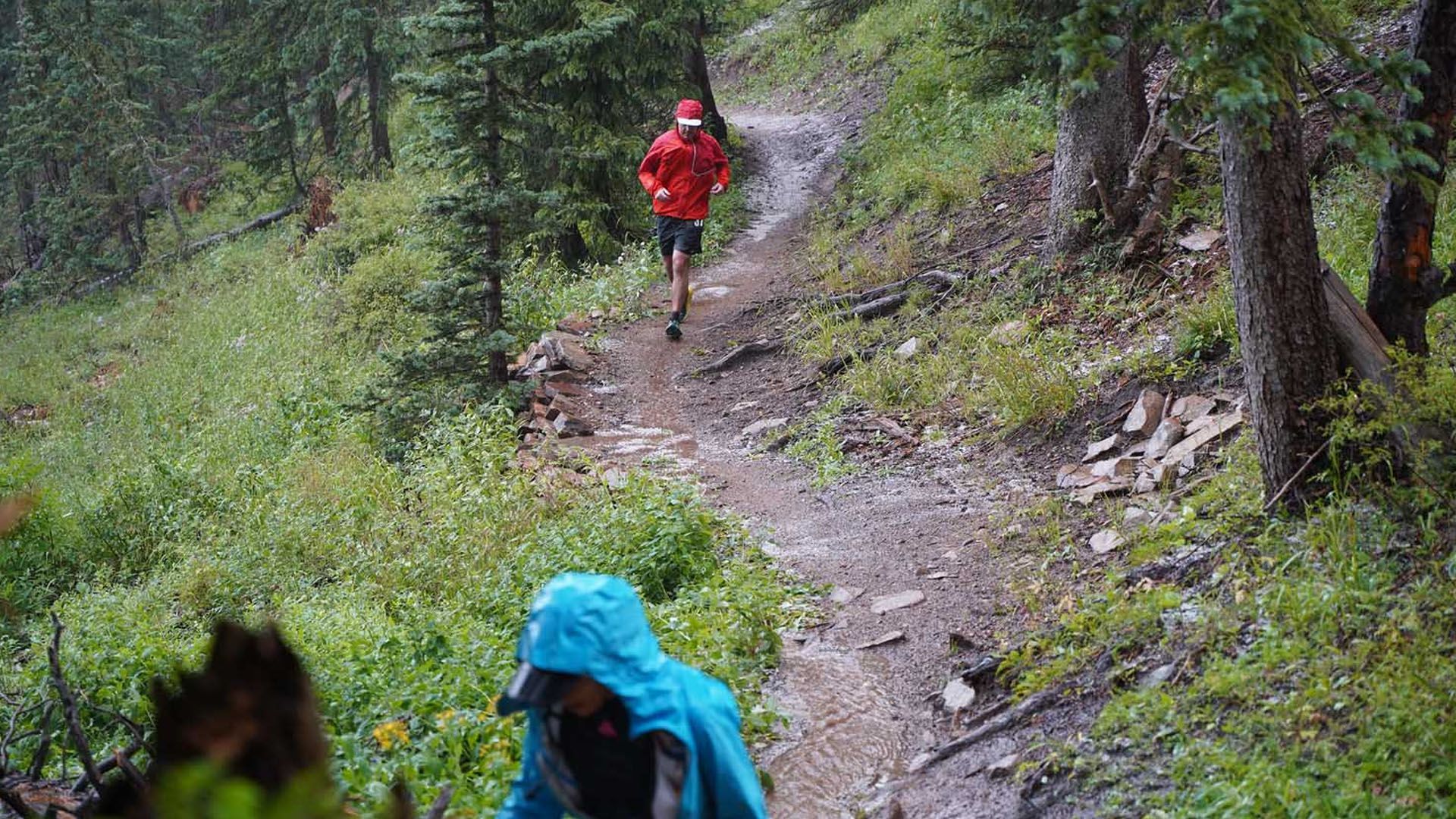 Two people in rain gear running on a muddy forest trail.