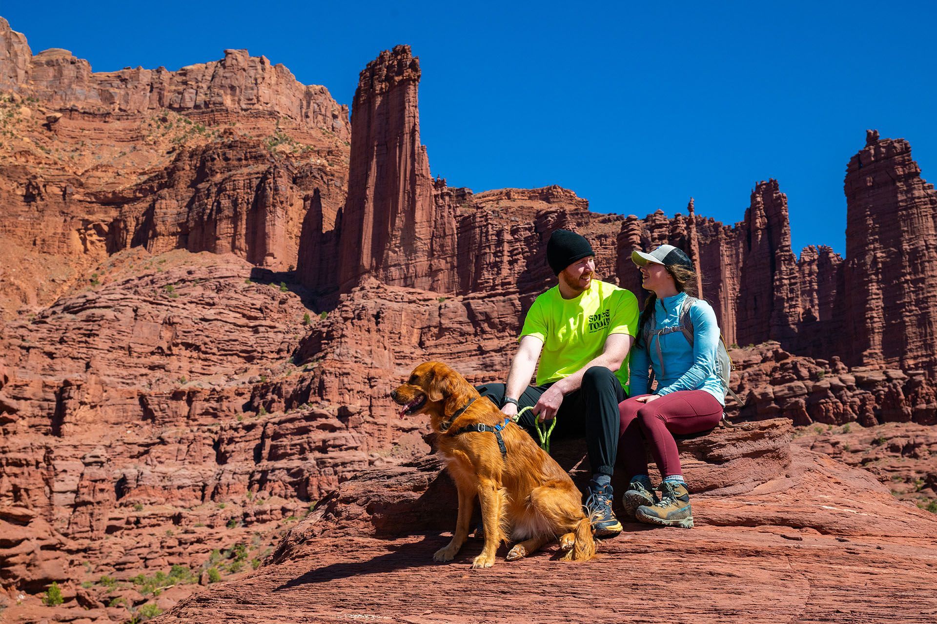 Two hikers sit on a rock with their dog in front of sandstone spires knows as Fisher Towers.