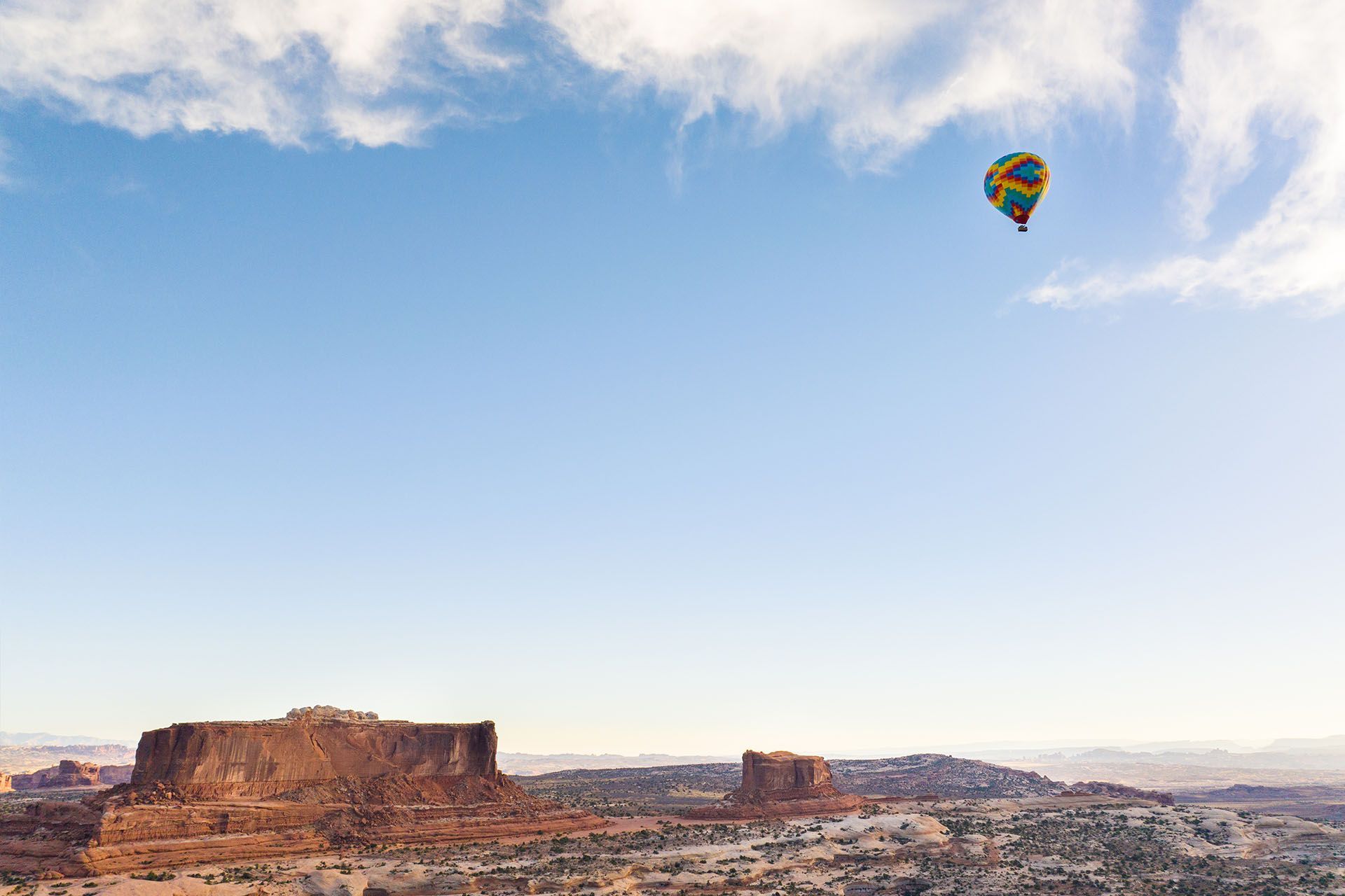 Hot air balloon over Monitor and Merrimac buttes in a desert landscape, against a blue sky with clouds.