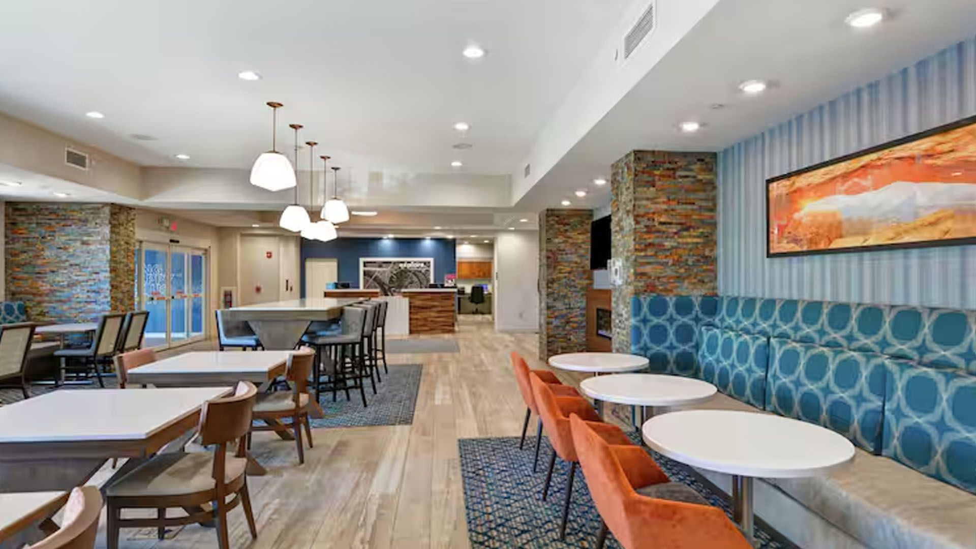 Hotel lobby with seating areas; blue, orange, and neutral tones; tables, chairs, and banquettes.