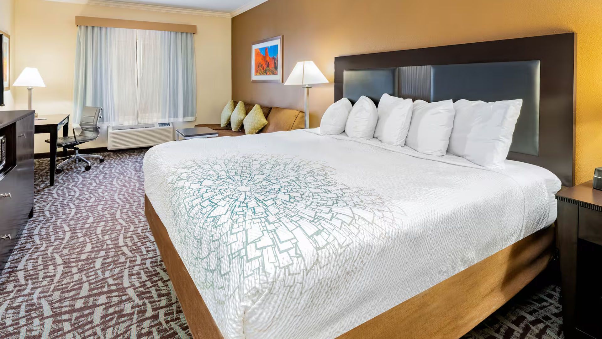 Hotel room with king-sized bed, desk, couch, and artwork. Light brown walls, patterned carpet.
