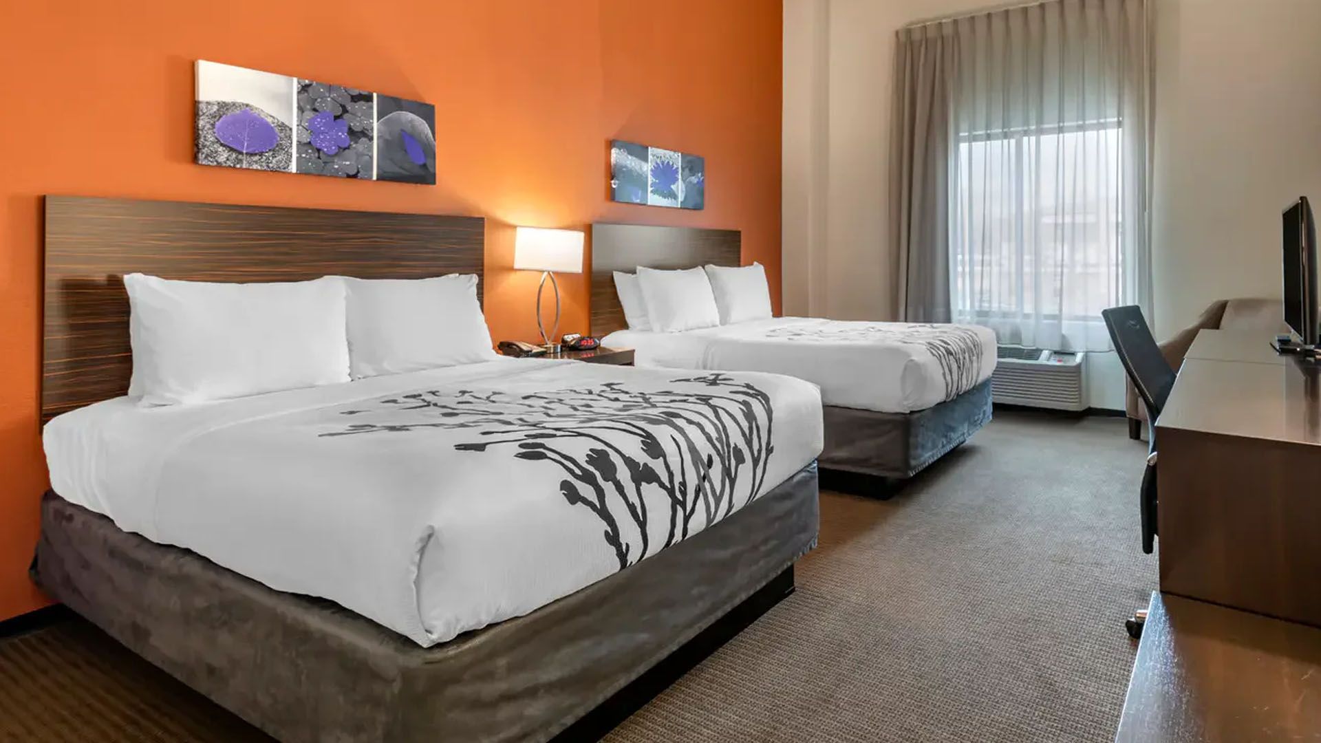 Two beds in a hotel room, orange wall, art, window with curtains, desk, chair, and TV.