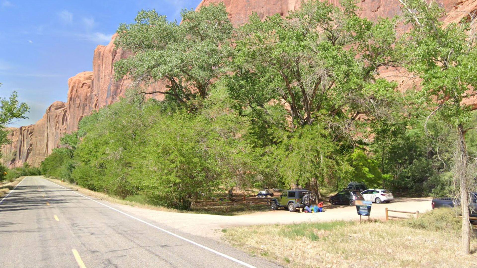 Roadside campsite with trees and red rock cliffs. Cars parked near a picnic area.