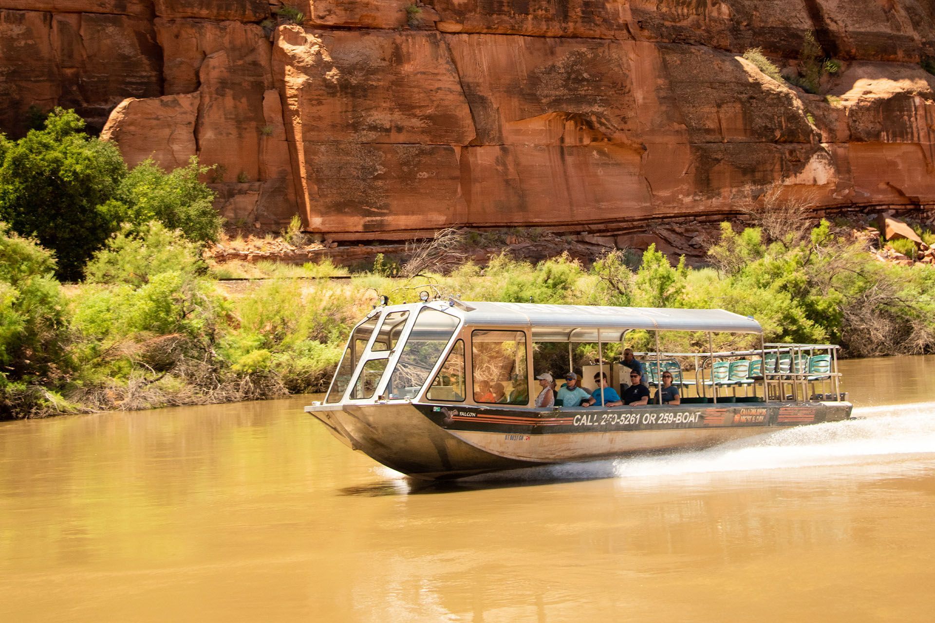 Jet boat with passengers on a brown river, canyon walls in the background. Sunny day.