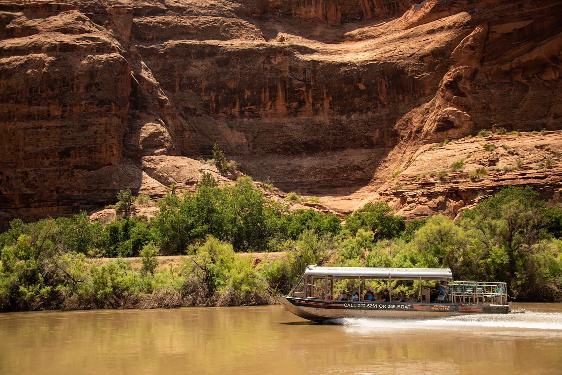 A boat with passengers cruises on a muddy river, canyon walls in the background.