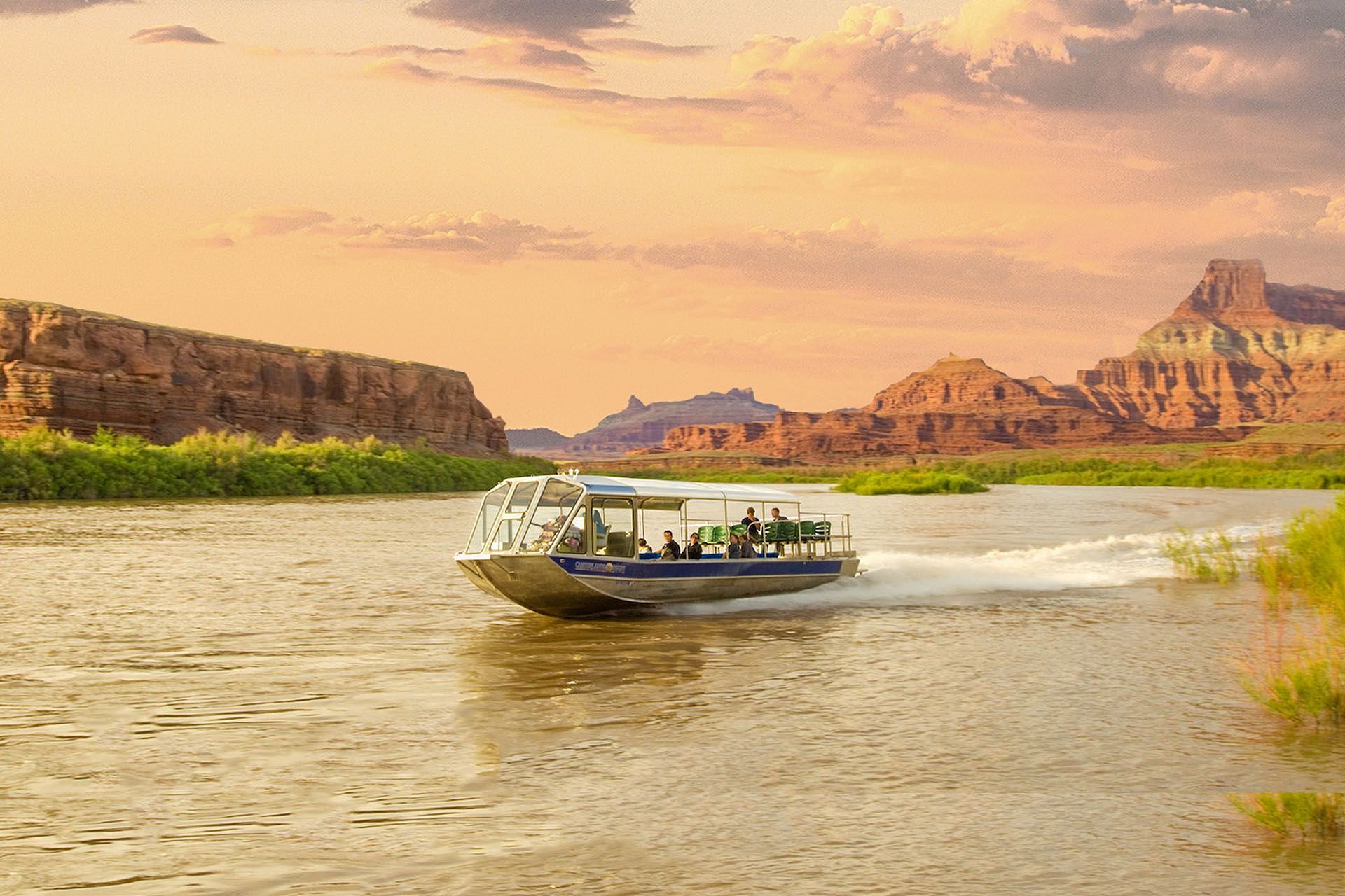 Boat on a river, passengers visible, against a backdrop of red rock cliffs at sunset.