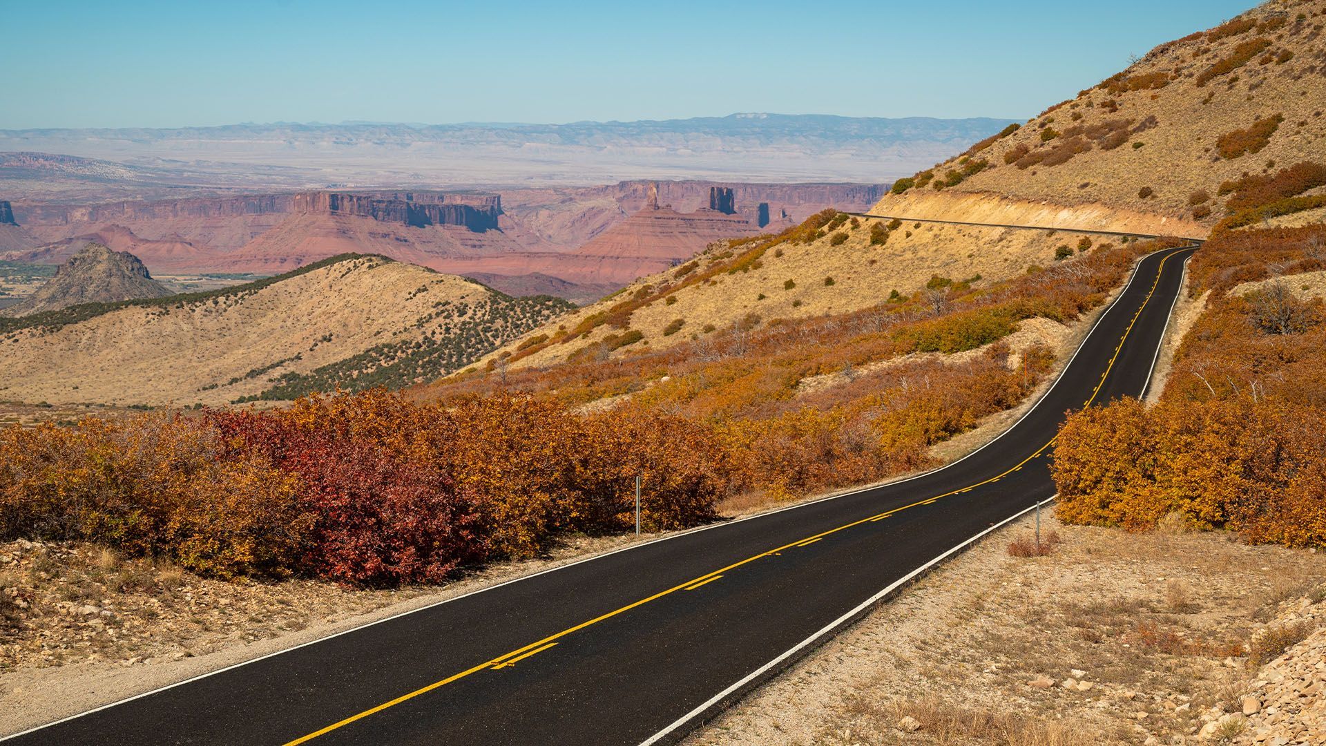 A winding asphalt road through a desert landscape with red rock formations and a clear blue sky.