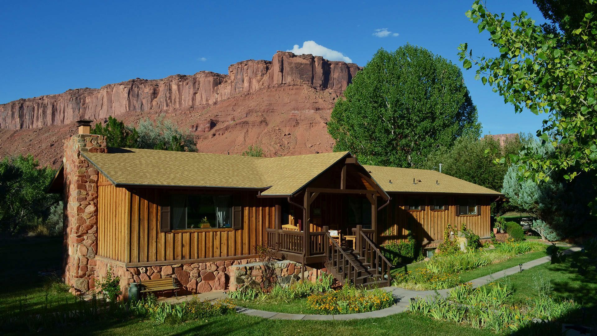 Rustic cabin nestled in a green yard with red rock mountains in the background under a blue sky.