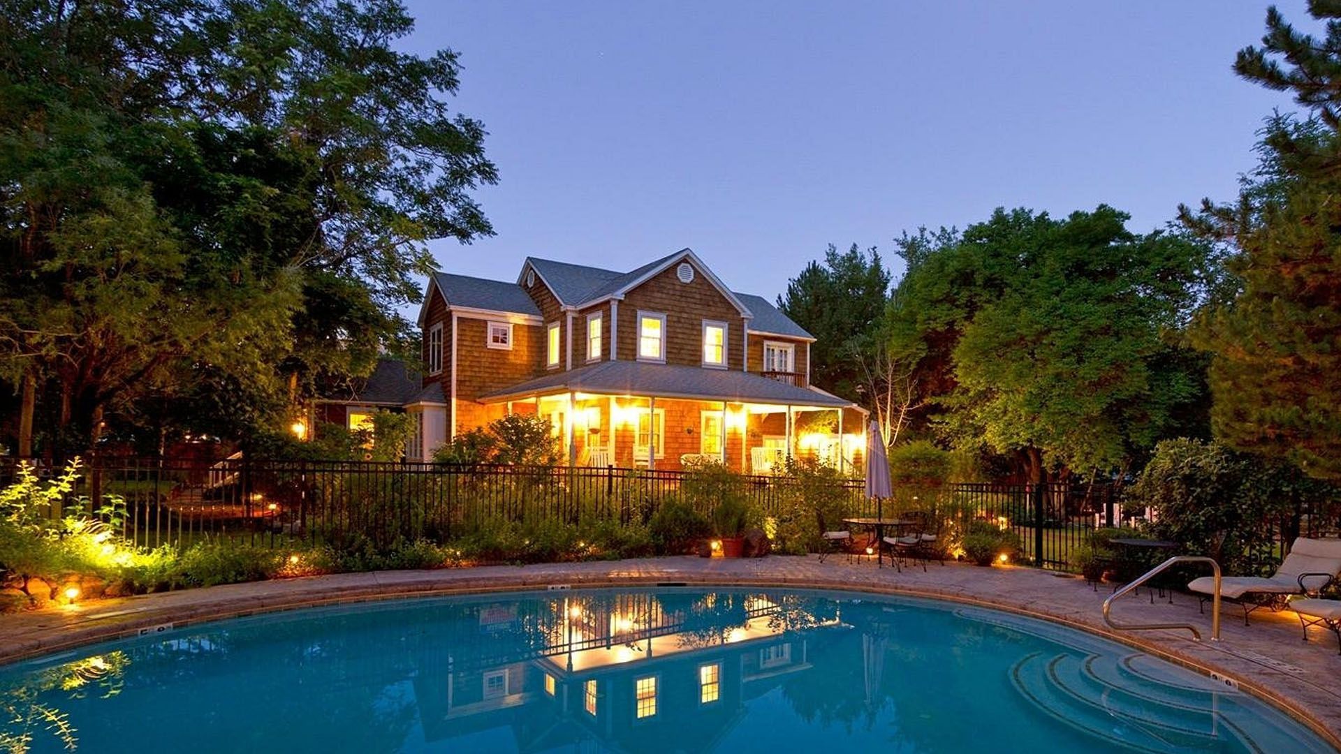 House with a pool at dusk, lit with warm lights. Lush trees surround the property.