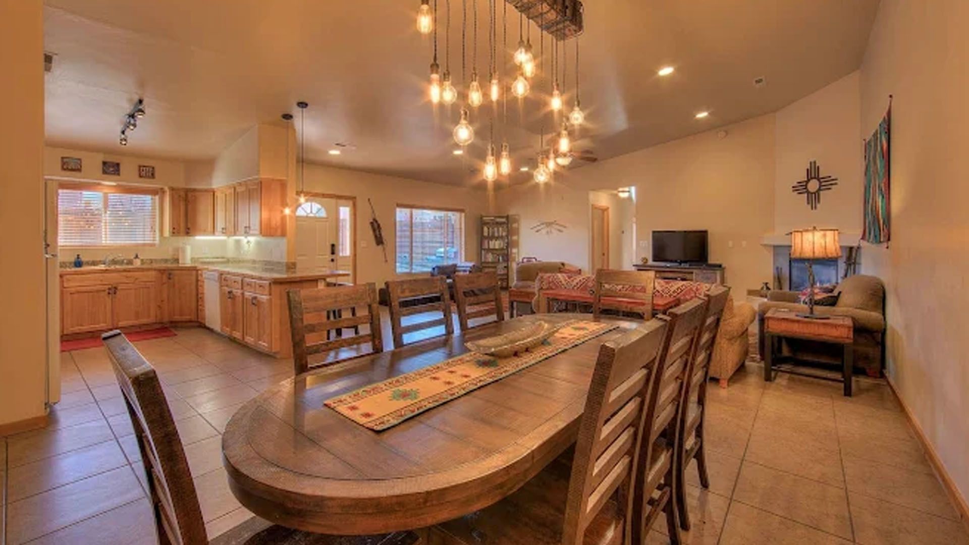Open-concept dining and living area. Long wooden table with chairs, kitchen in the background, warm lighting.