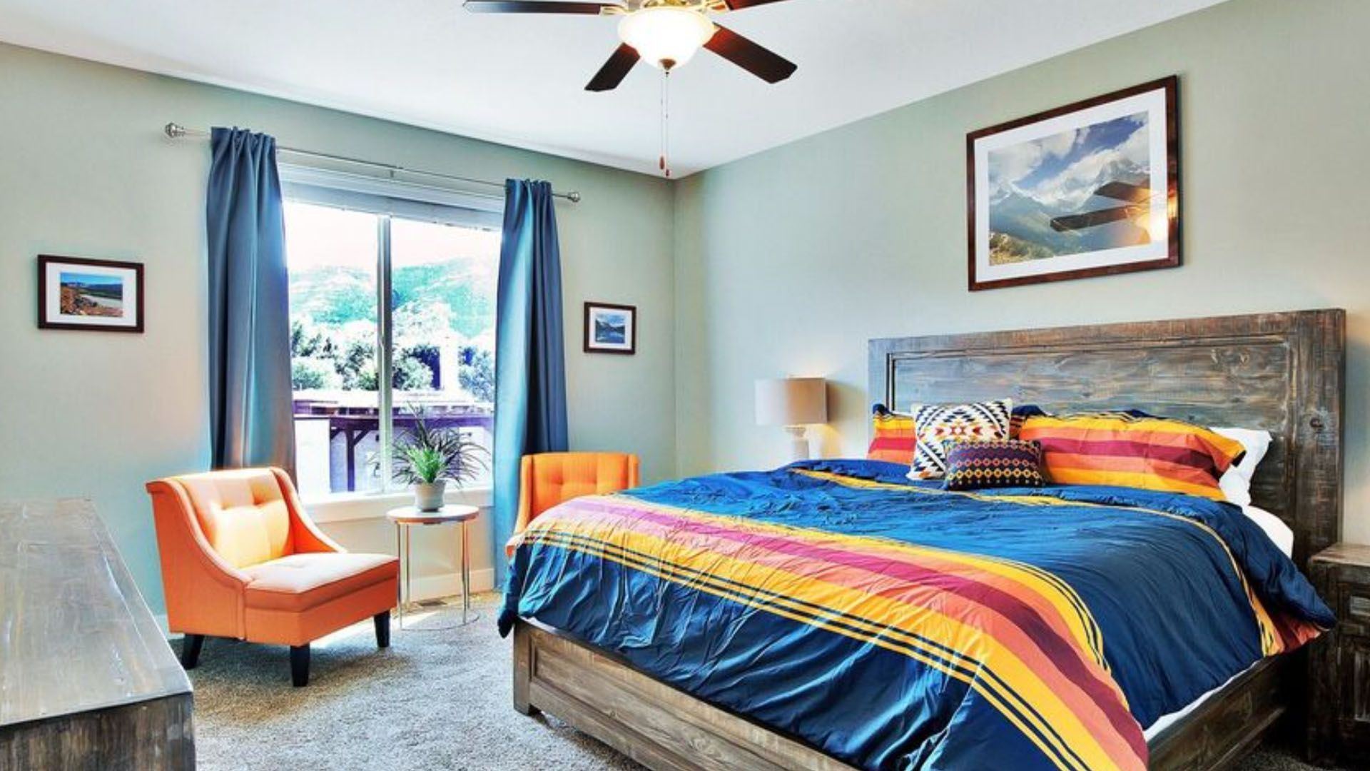 Vacation rental bedroom with bed, orange chairs, blue curtains, and landscape art.