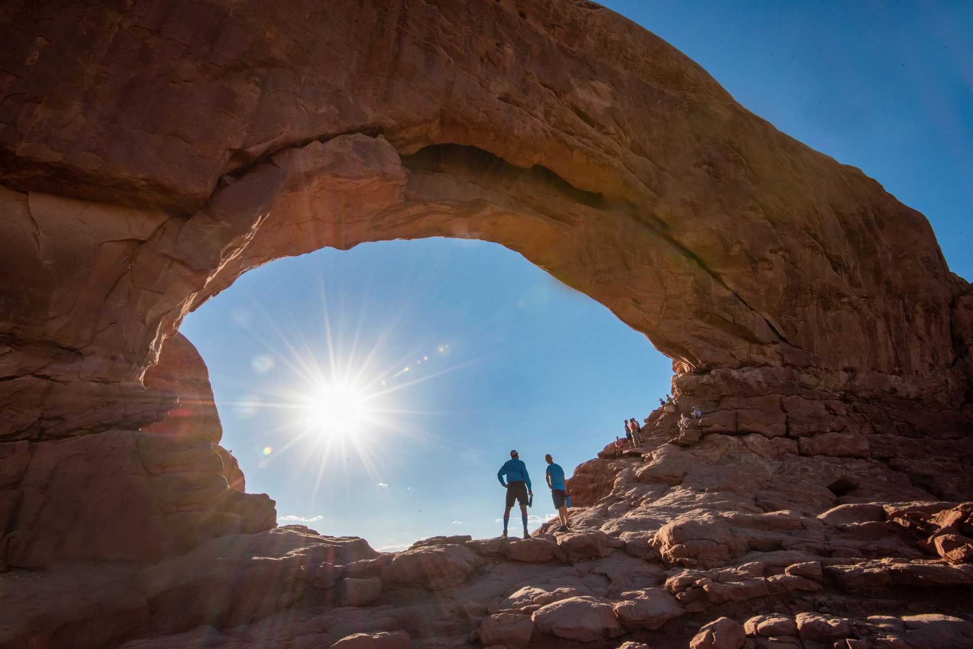 Two figures stand under a large rock arch, sunburst in background, blue sky.