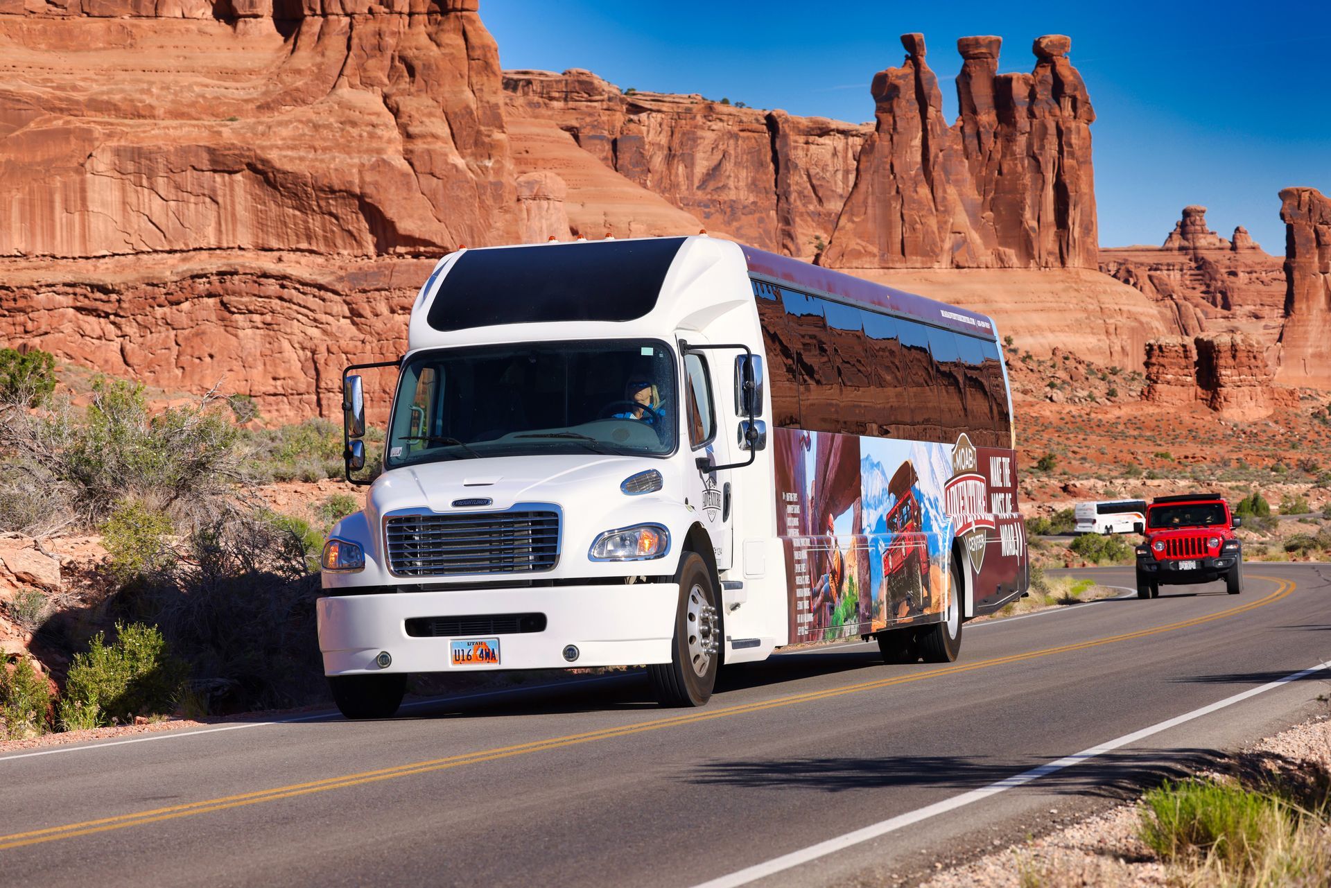 White tour bus on road in front of red rock formations, small red SUV in background.