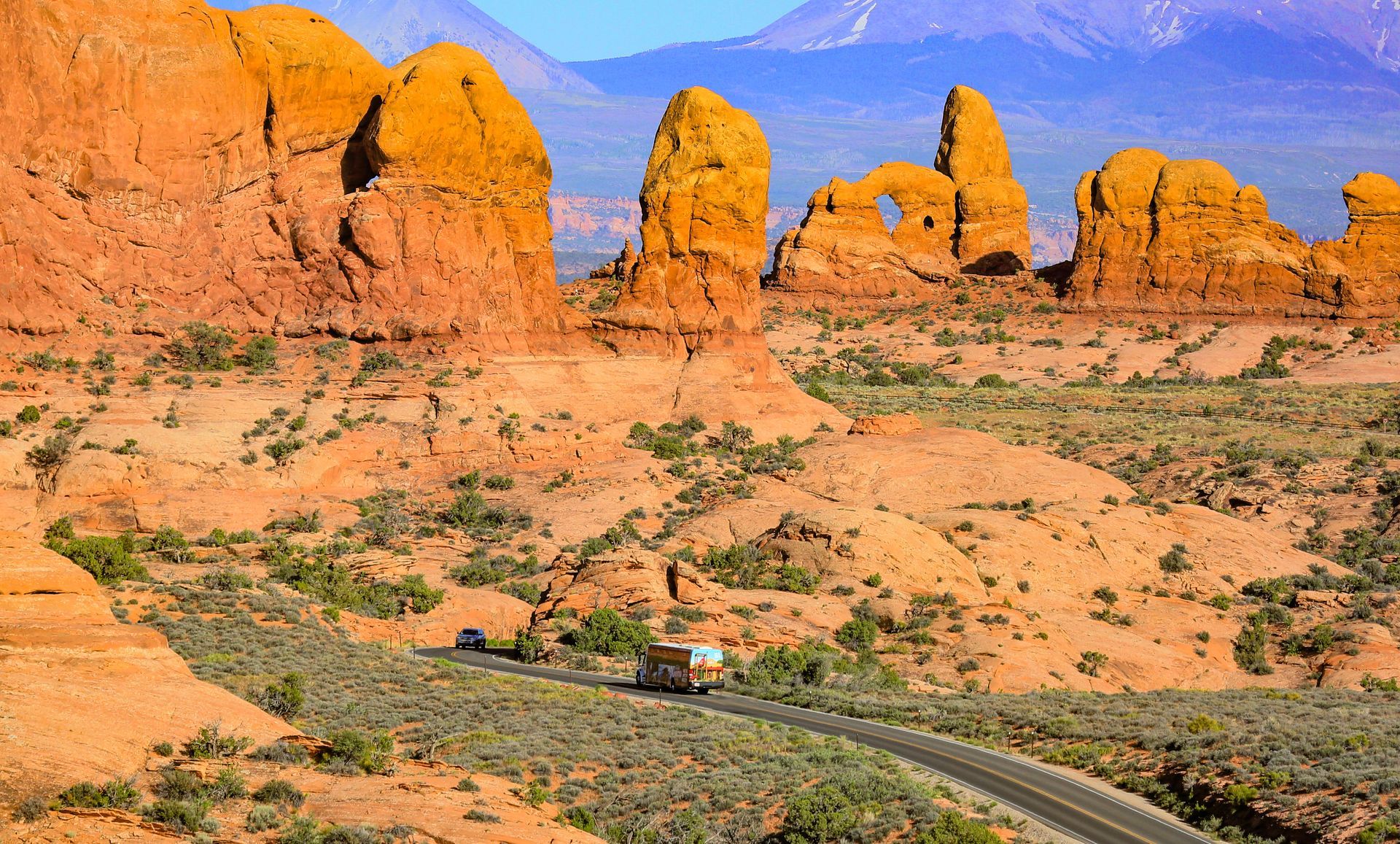 Desert landscape with sandstone formations, road, and vehicle; mountains in the background.