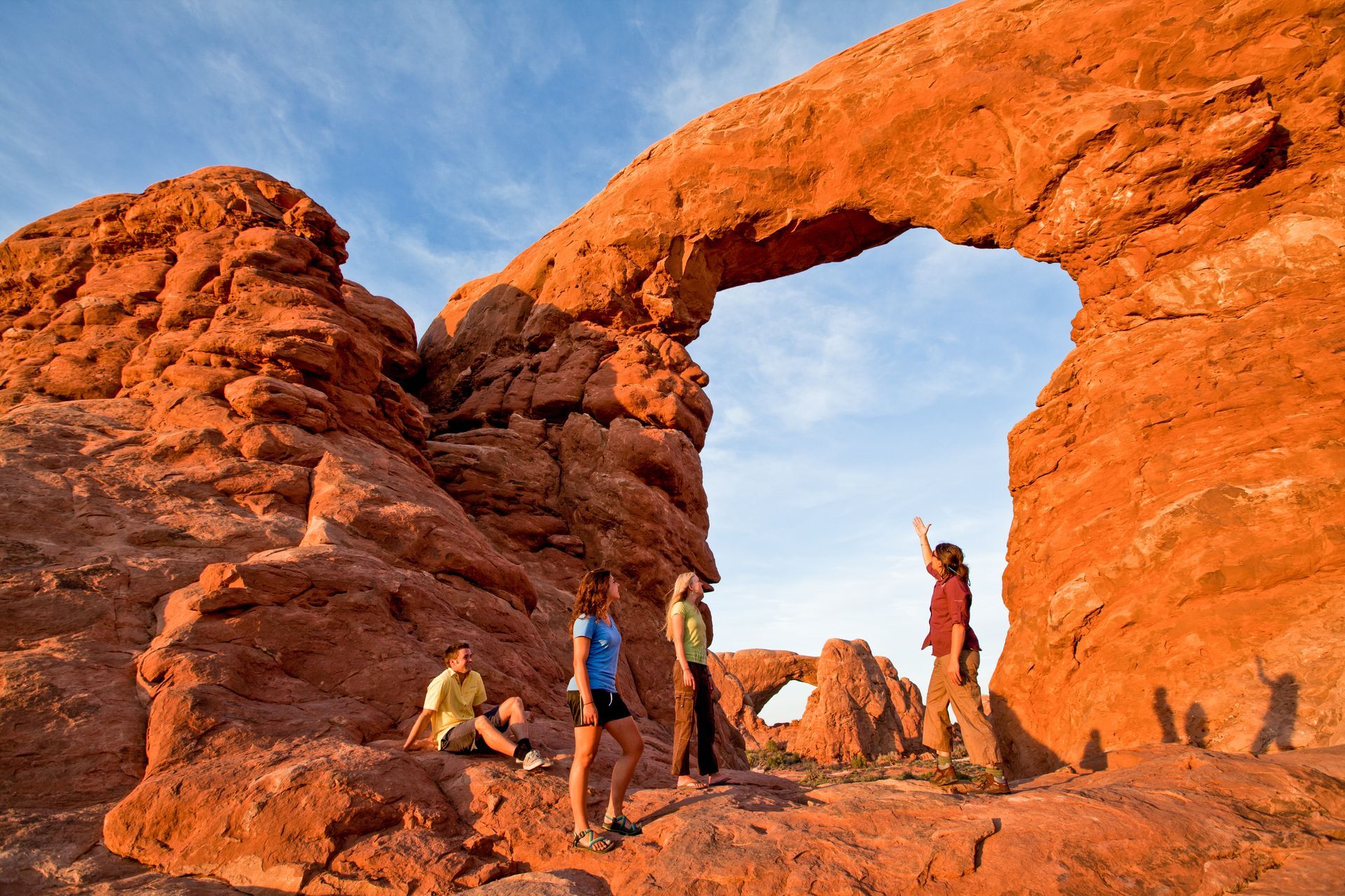 People standing beneath a large, red rock arch in a desert setting, with blue sky above.