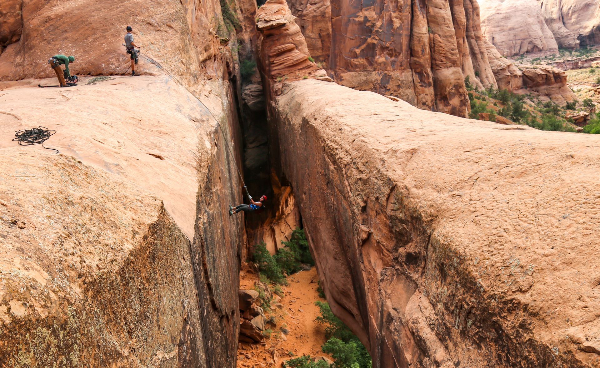 A climber rappels down a narrow slot canyon between red rock walls; two figures stand at the top.