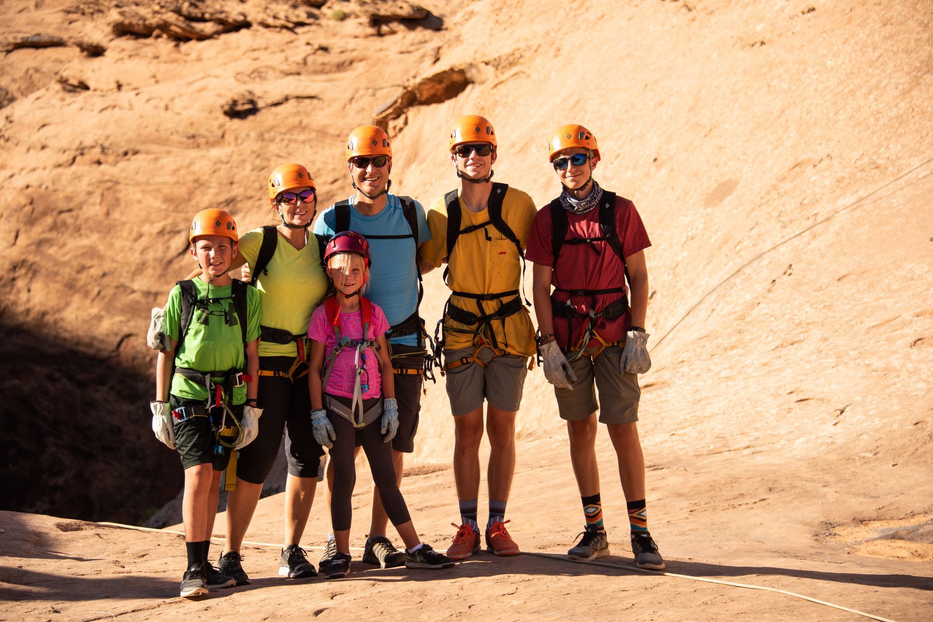 Family of six in climbing gear posing in front of a sandstone cliff.