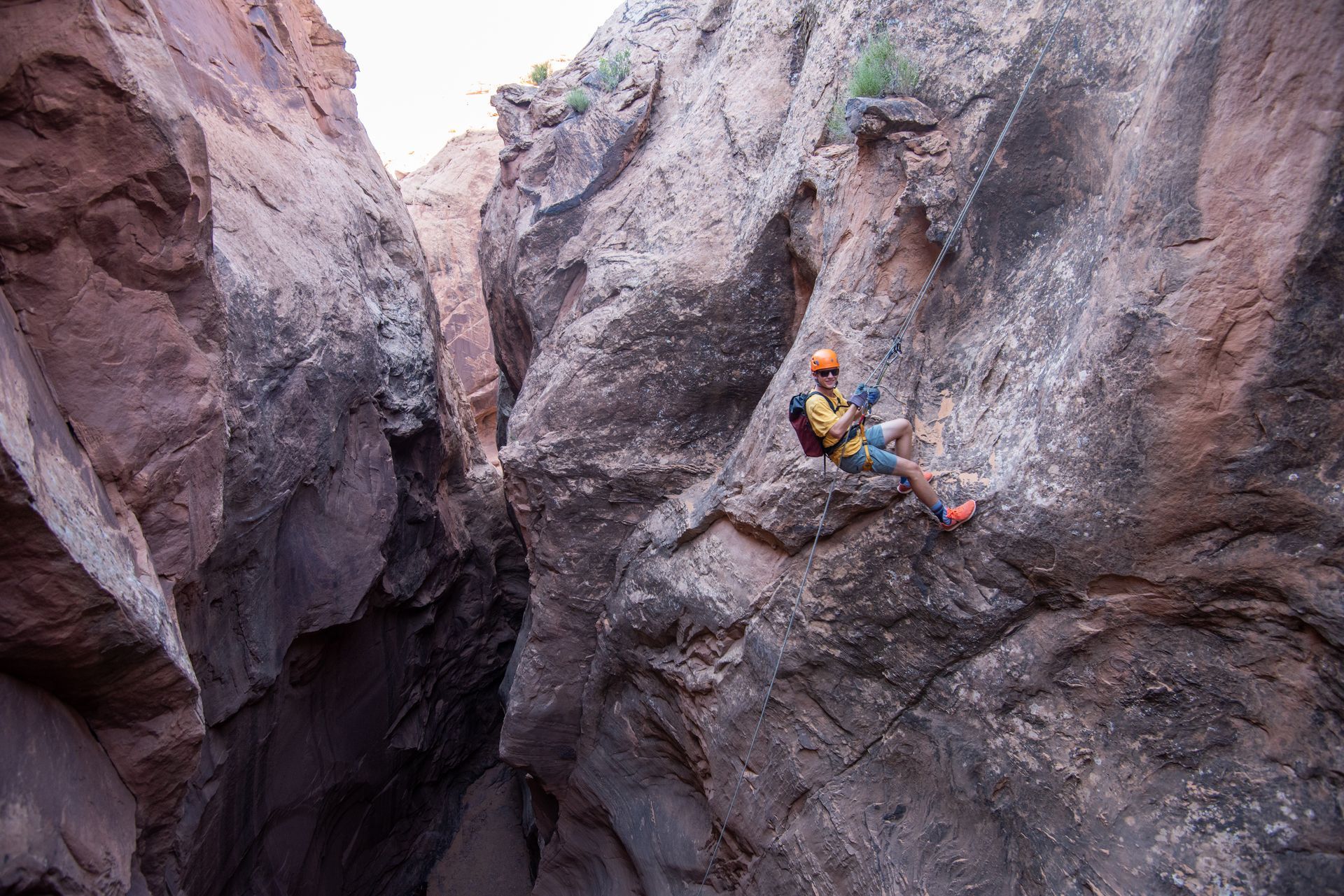 Man rappels down a narrow canyon, wearing a helmet and harness, against a reddish-brown rock face.