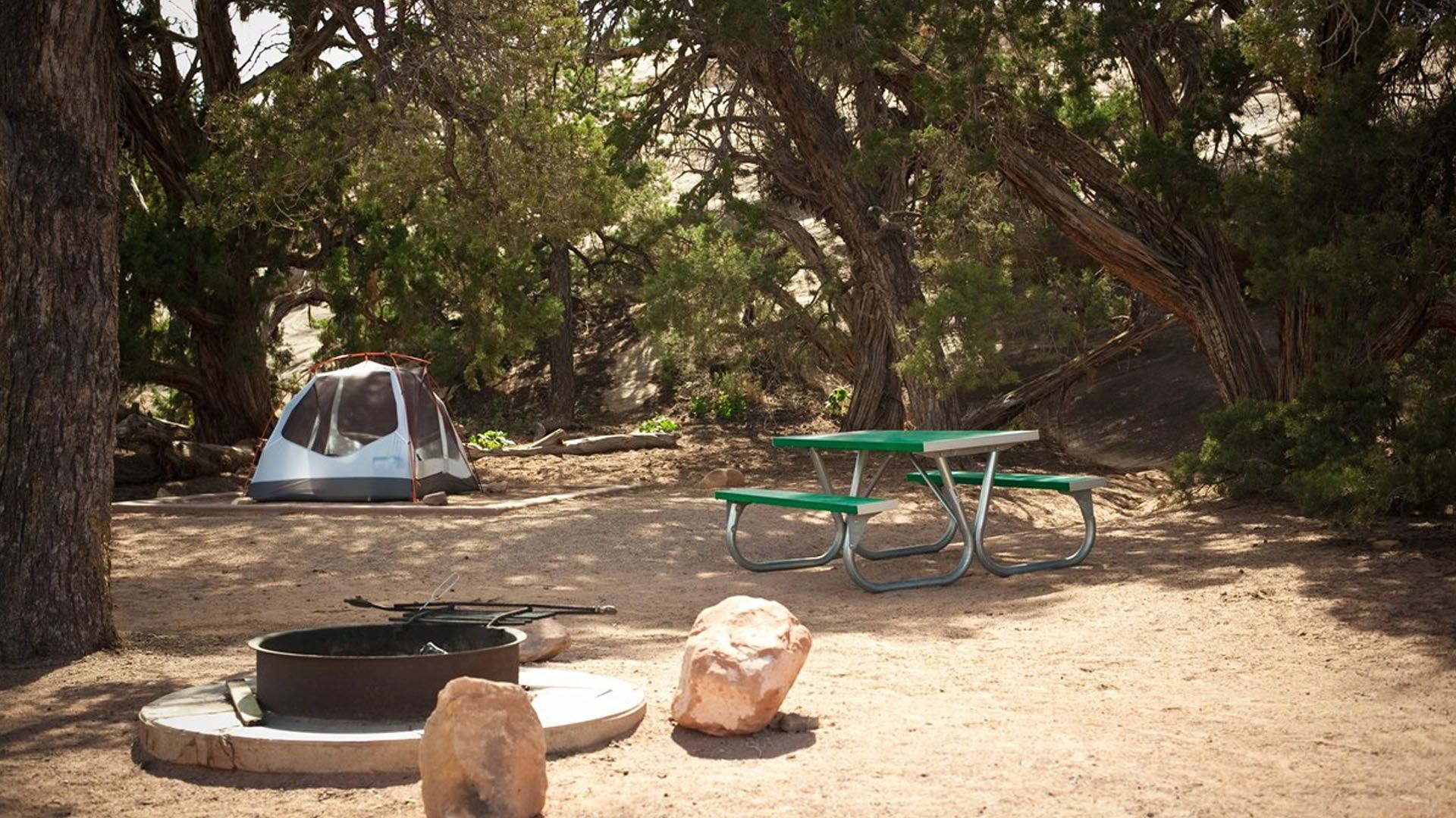 Camping scene: tent, picnic table, fire pit, and large rocks in a shaded area.