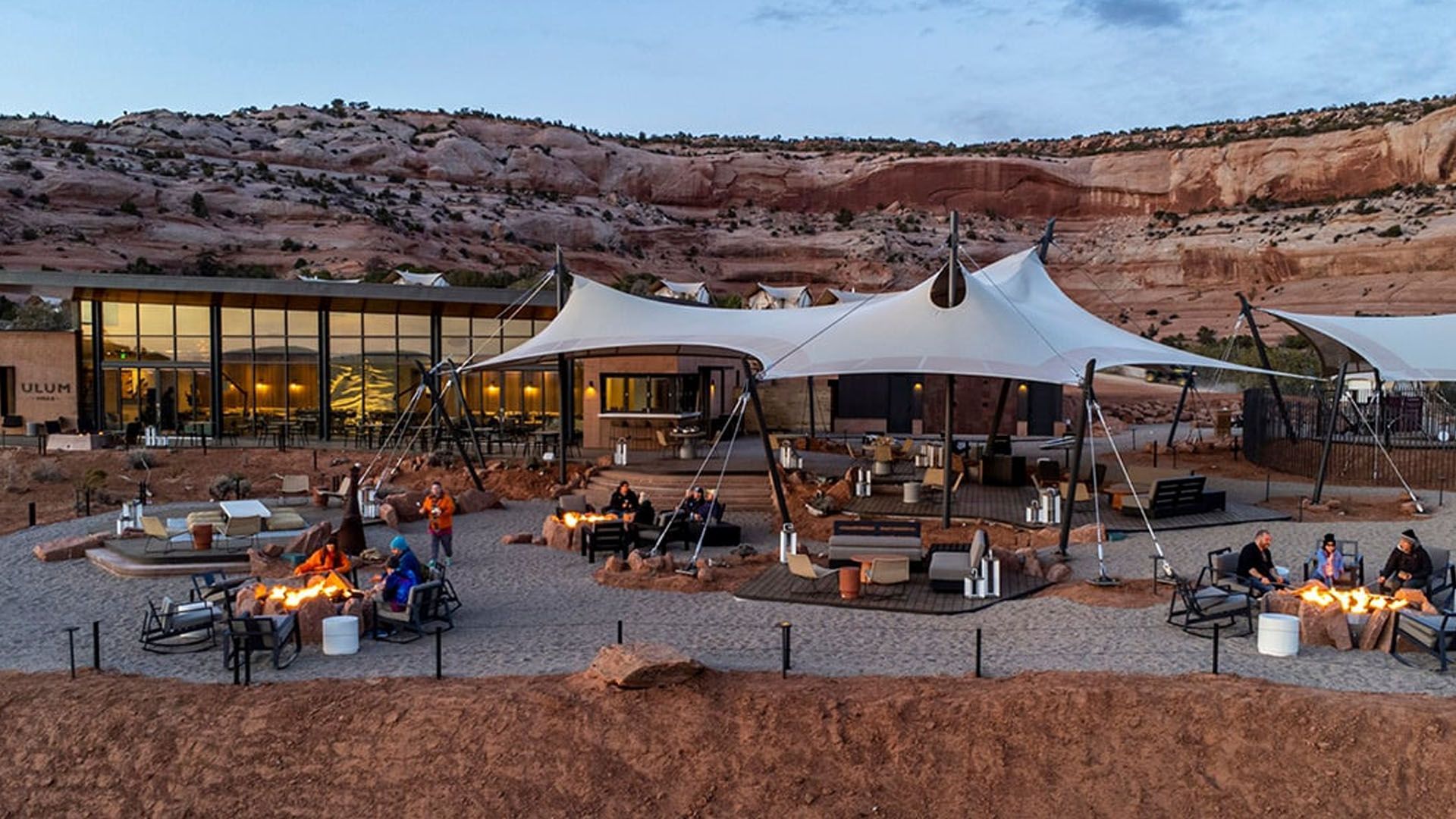 People gathered around fire pits under a large tent and near a building, in a desert landscape at dusk.