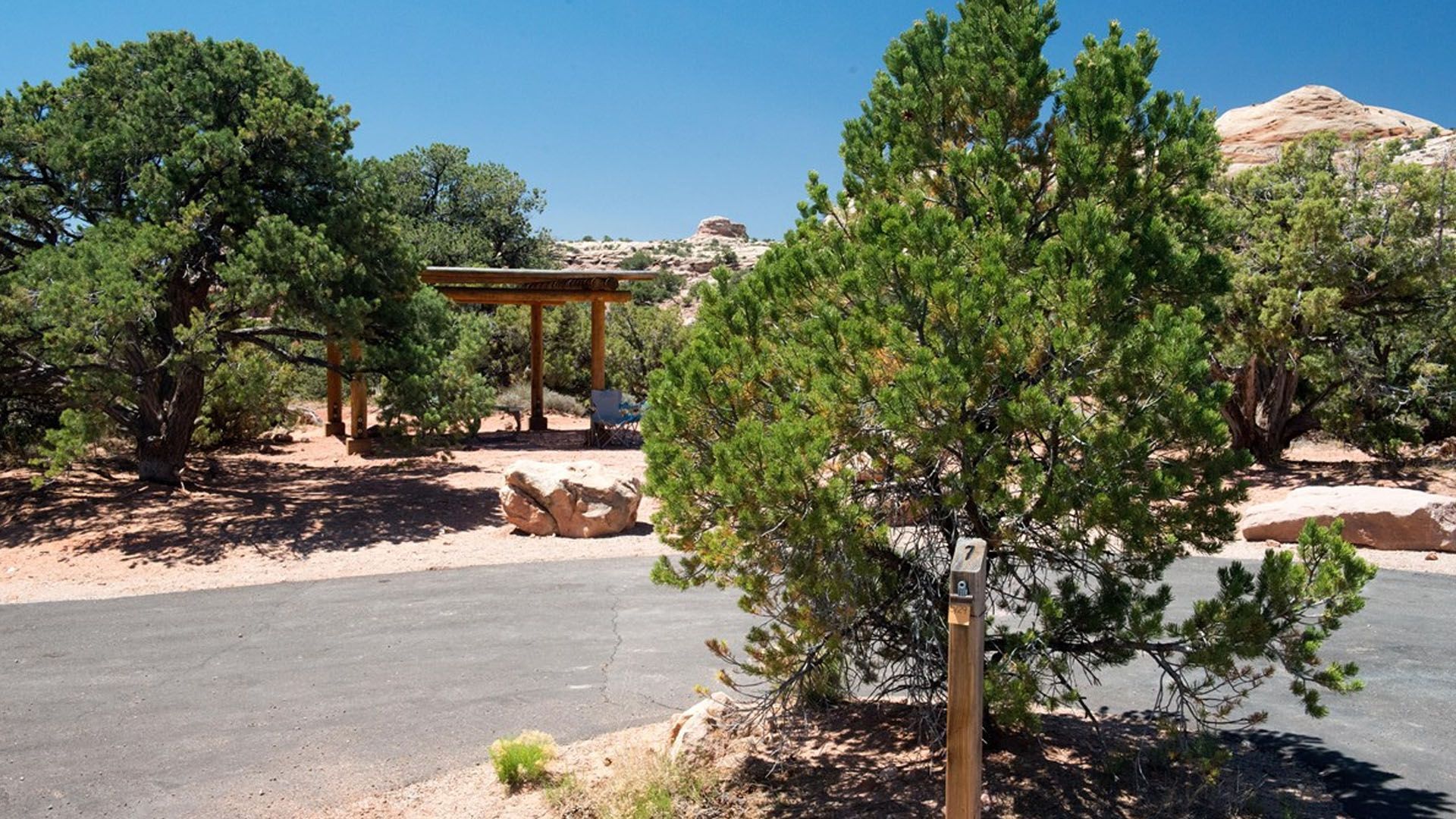 Paved road through campground. Trees surround a wooden shade structure at a campsite. Bright blue sky in background.