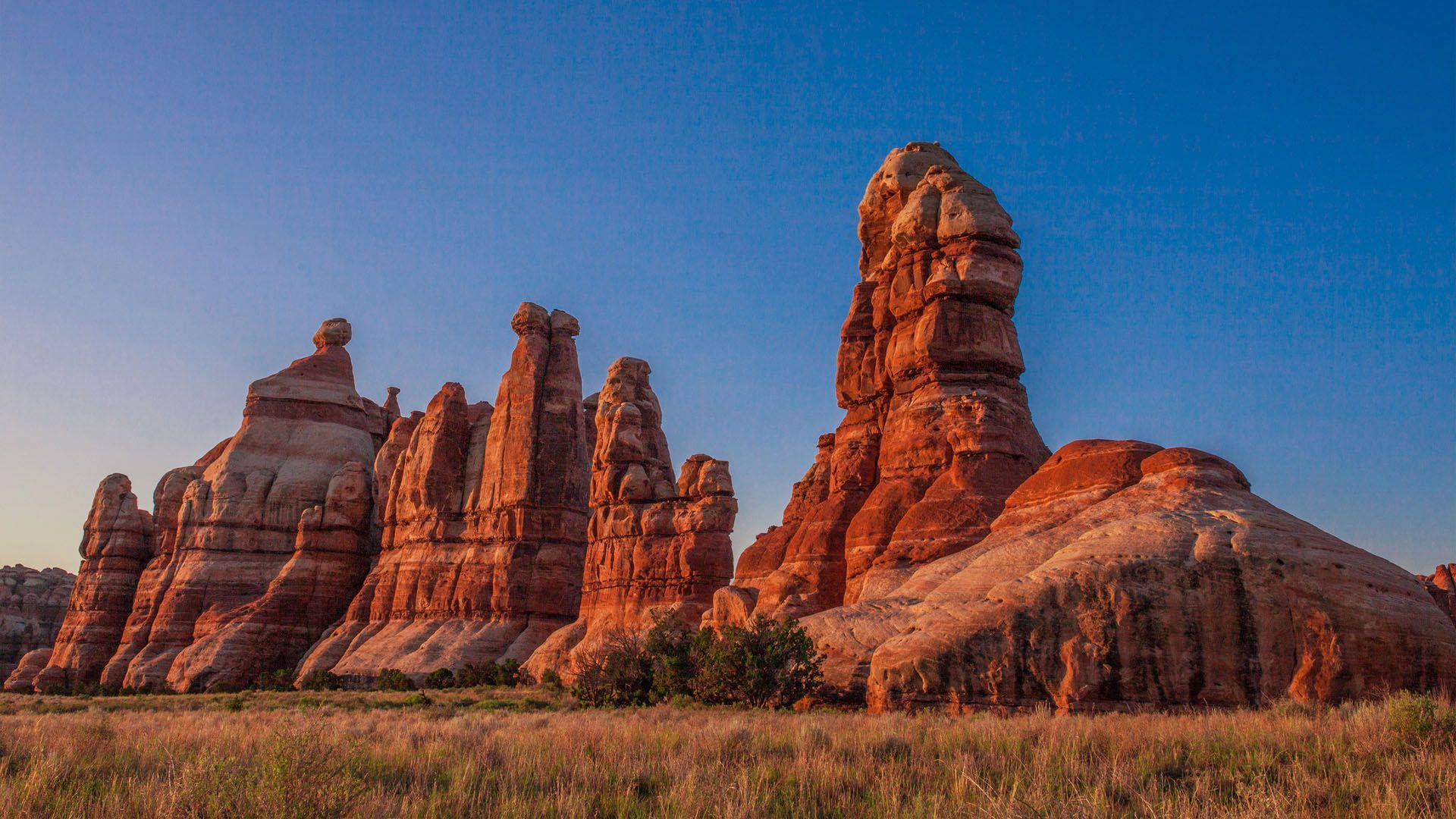 Red rock formations stand tall against a clear blue sky, grassy foreground.