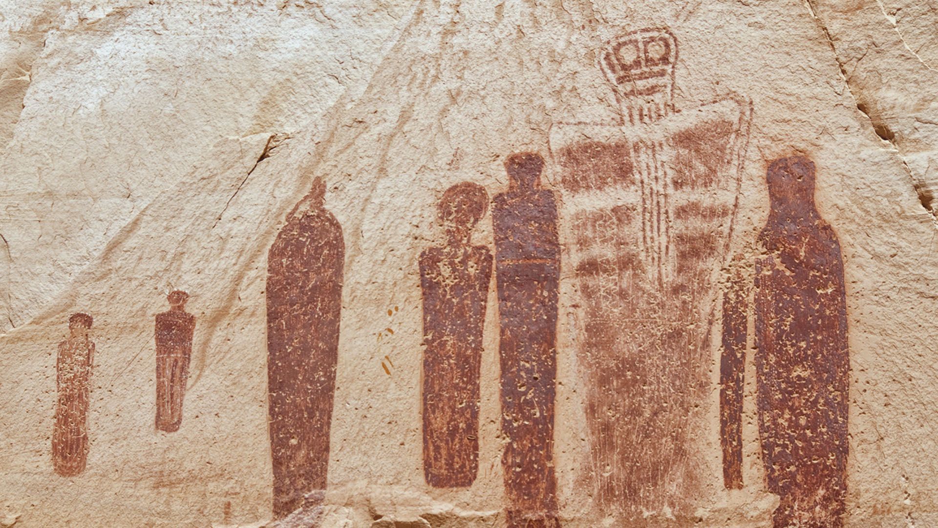 Red rock art figures on sandstone, depicting human-like forms in a line.