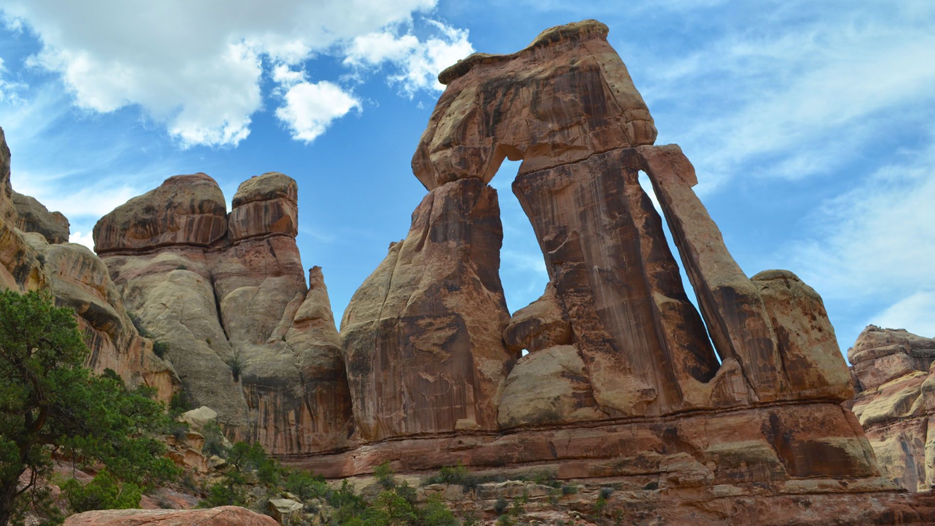 Canyonlands National Park: Brown sandstone rock formation with an arch against a blue sky with white clouds.