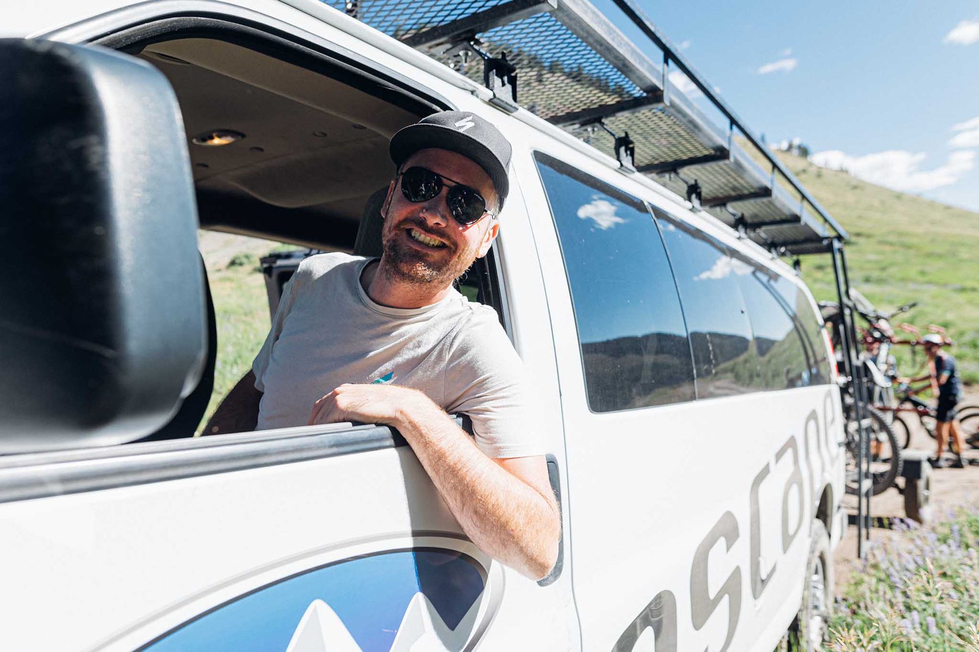 Man in sunglasses smiles from van window on a dirt road, mountain backdrop with mountain bikers gathered behind the van.