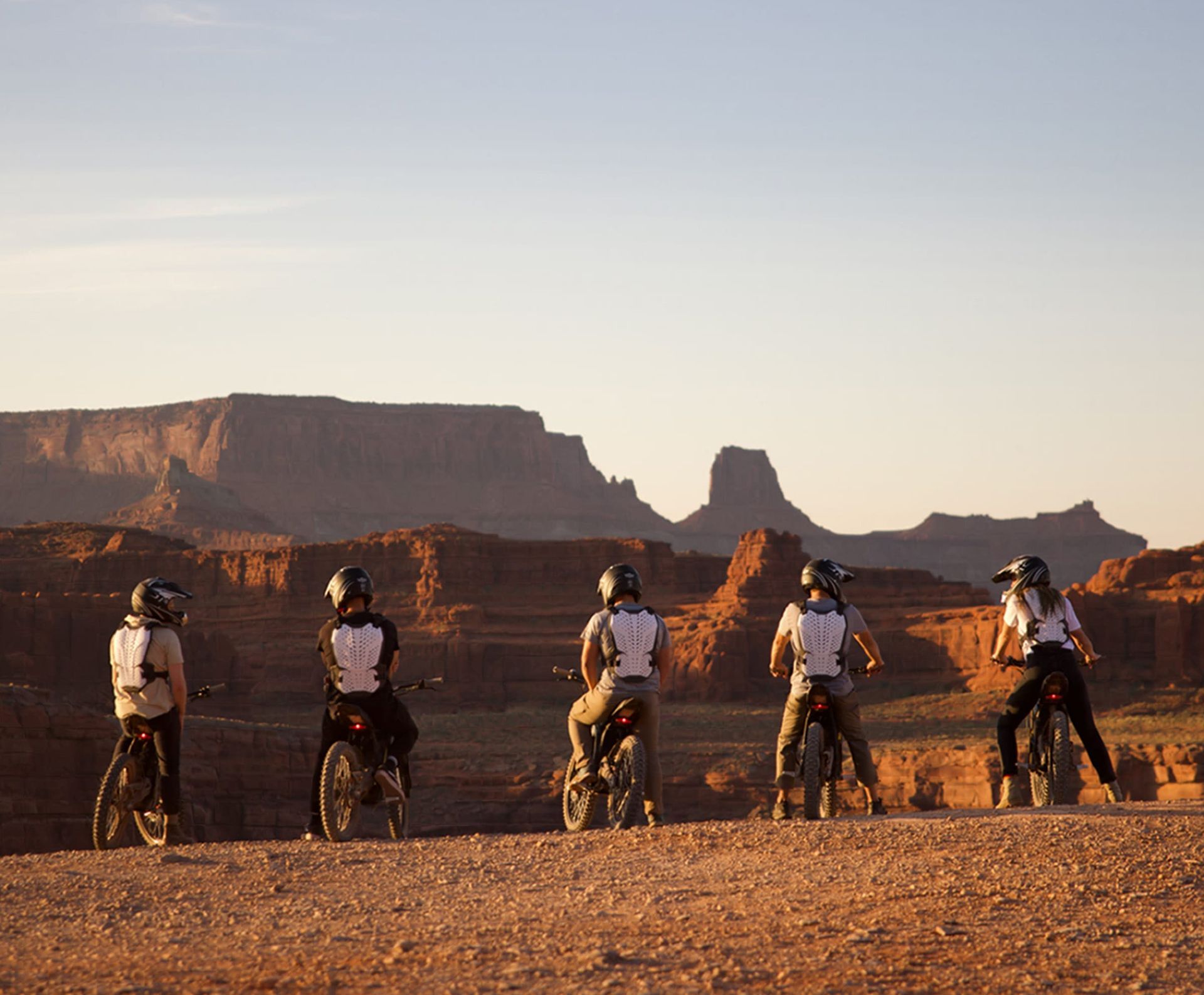 Five cyclists on e dirt bikes, facing a desert landscape at sunset.