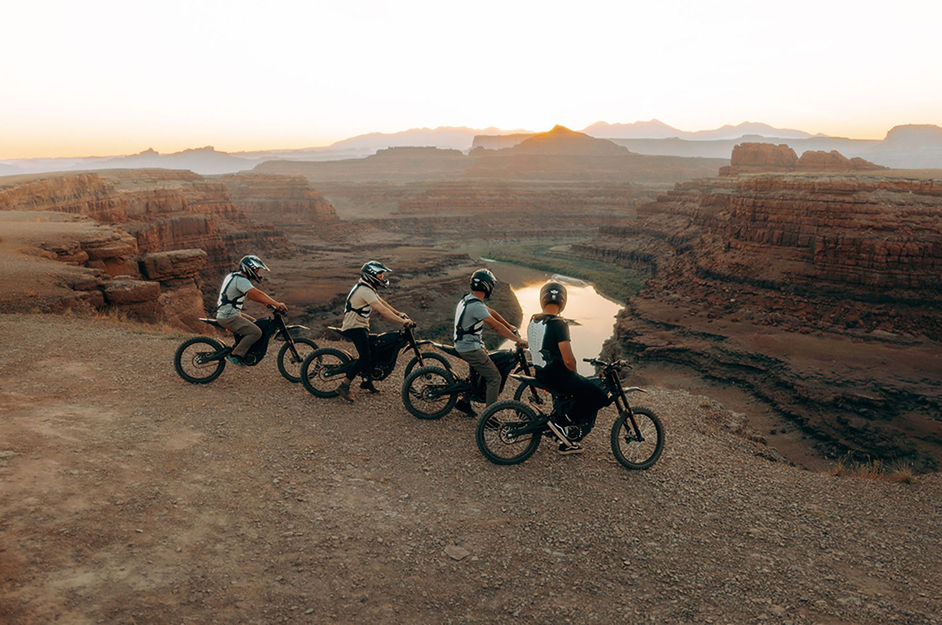 Four people on electric bikes atop a desert mesa at sunset, overlooking a river canyon.